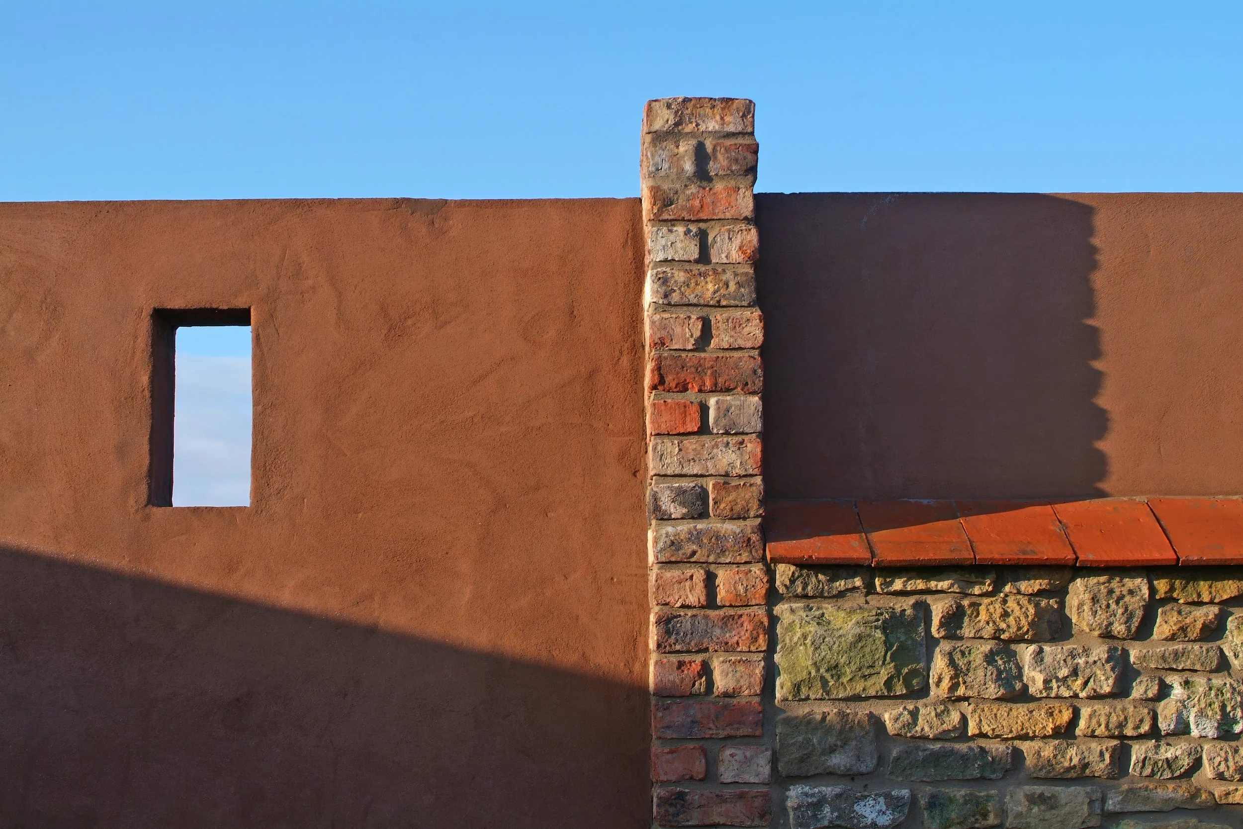 Close-up view of a wall with a small rectangular window on the left and a brick chimney in the center, with a blue sky background.