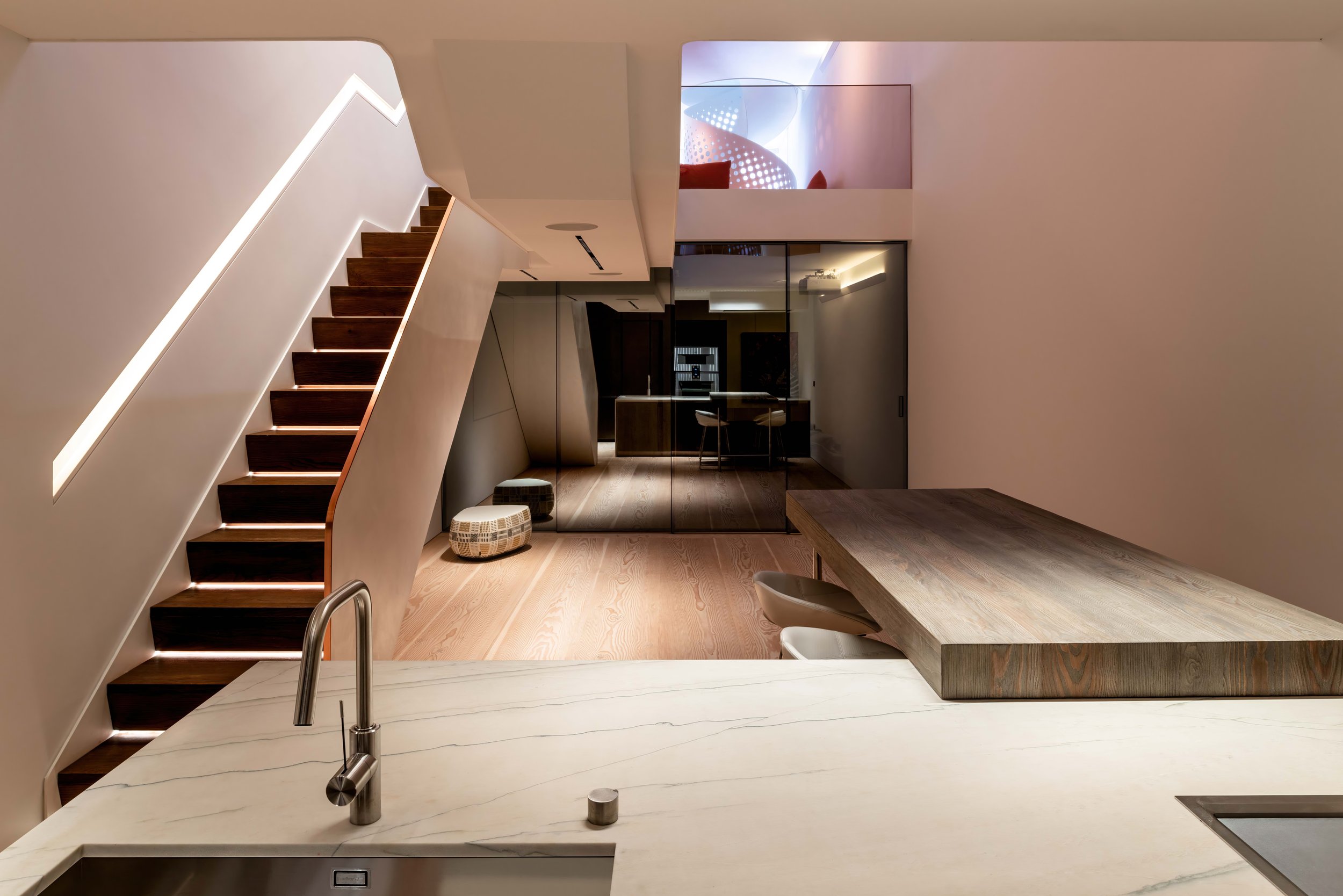 Modern interior with a staircase featuring illuminated steps, a glass-walled dining area, and a kitchenette with a marble countertop.