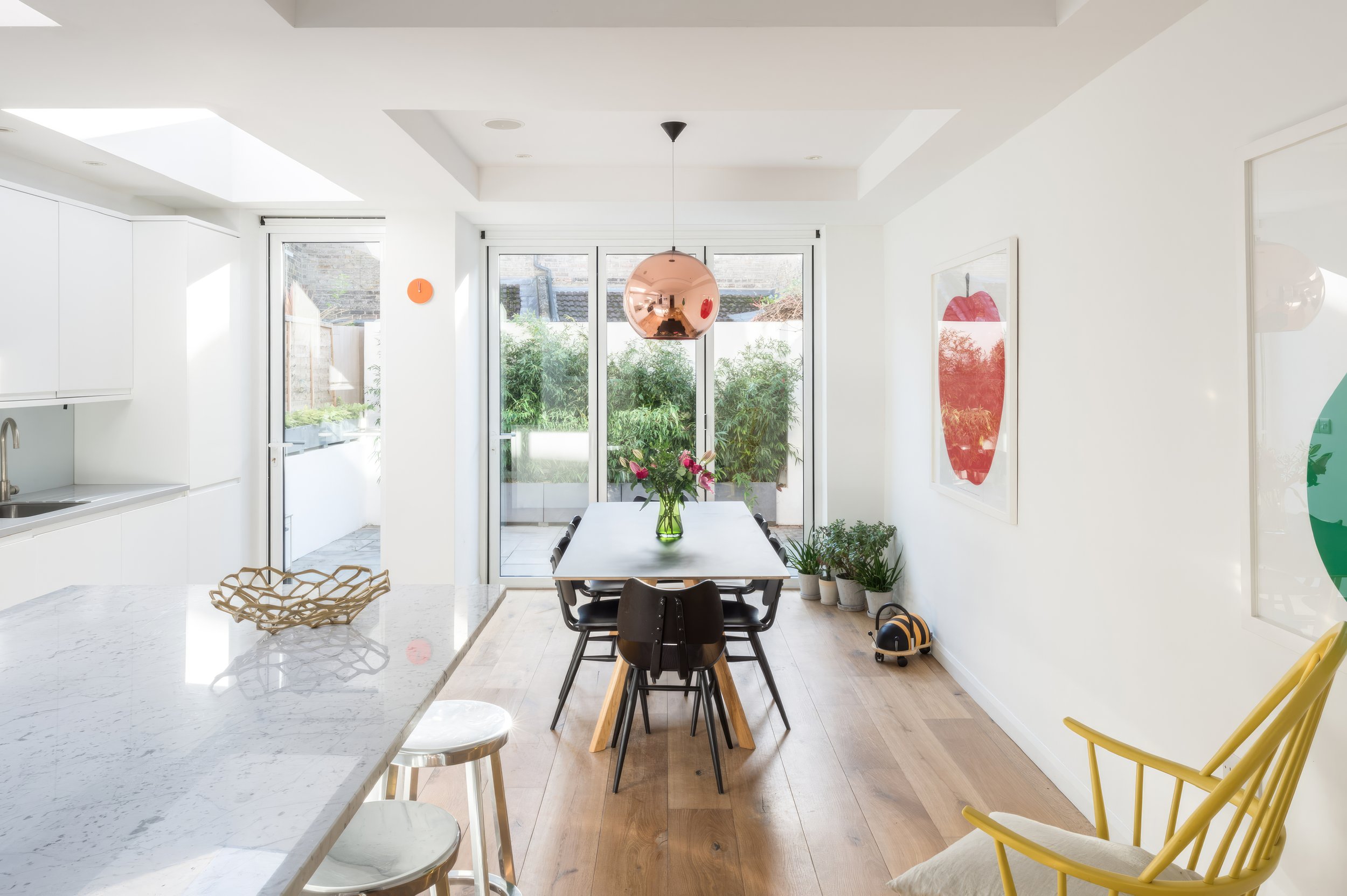 Bright, modern dining area with a white table, black chairs, and a vase of pink flowers. Glass sliding doors lead to an outdoor garden. Artwork of a red apple hangs on the wall, and a yellow chair is partially visible in the foreground.