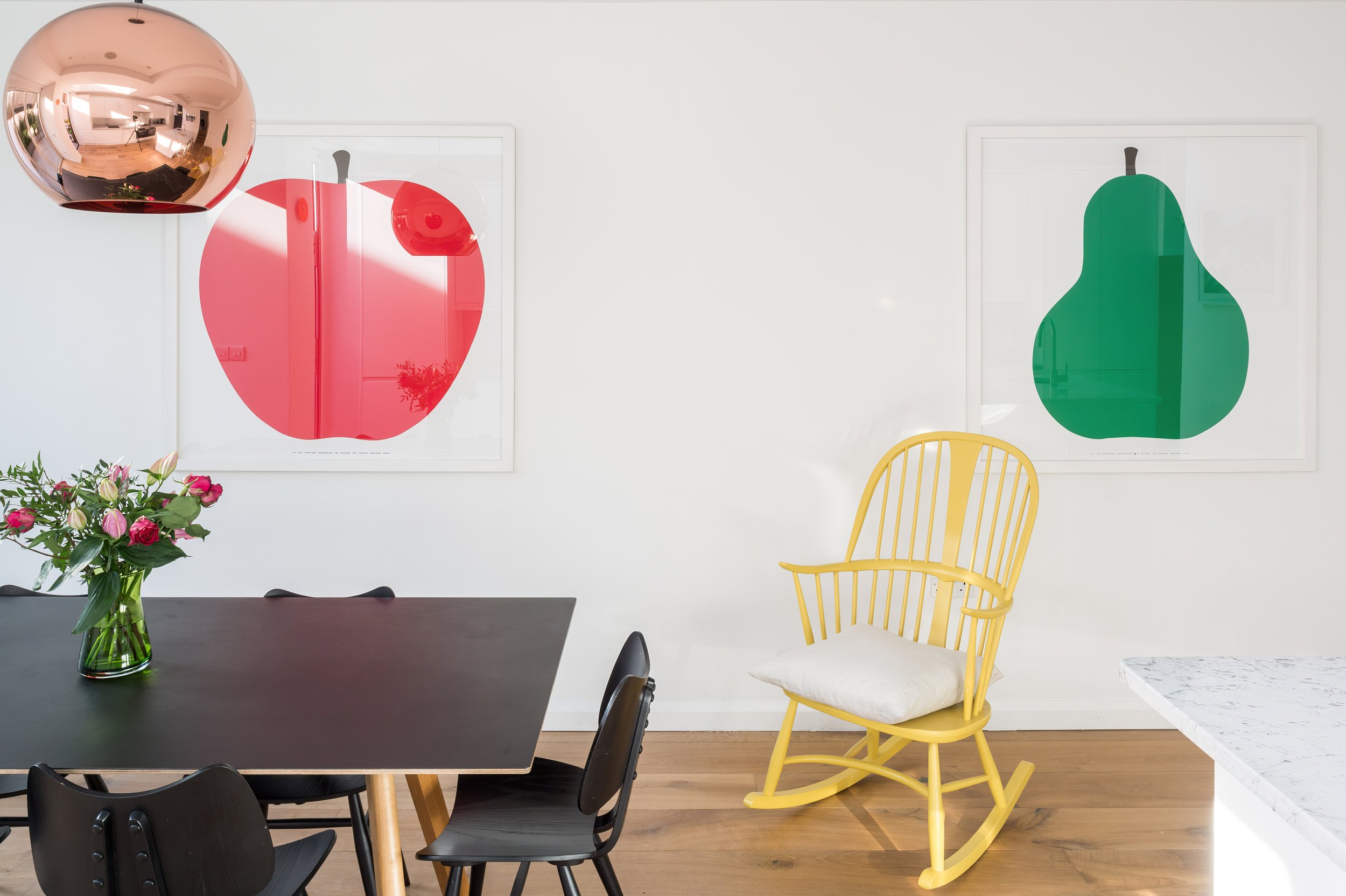A dining room with a black table, black chairs, a yellow rocking chair with a white cushion, a vase of pink and white flowers, and two large framed art prints of colorful apples and pears on the white wall.
