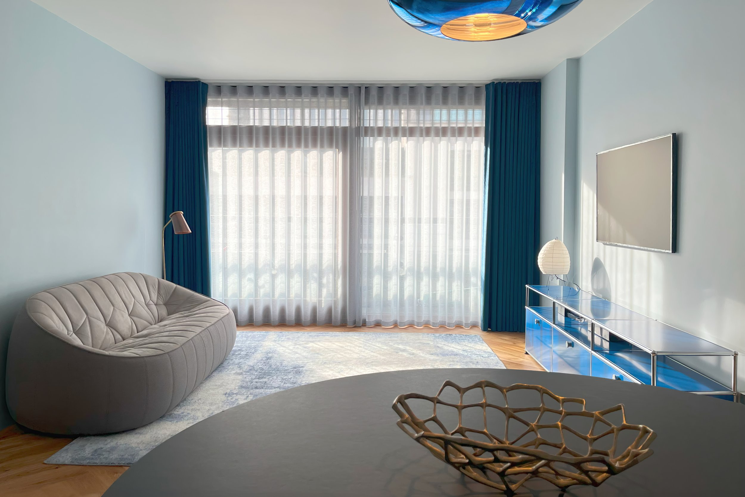 Modern living room with large windows, blue curtains, a beige couch, a blue TV stand, a wall-mounted TV, and a circular ceiling light.