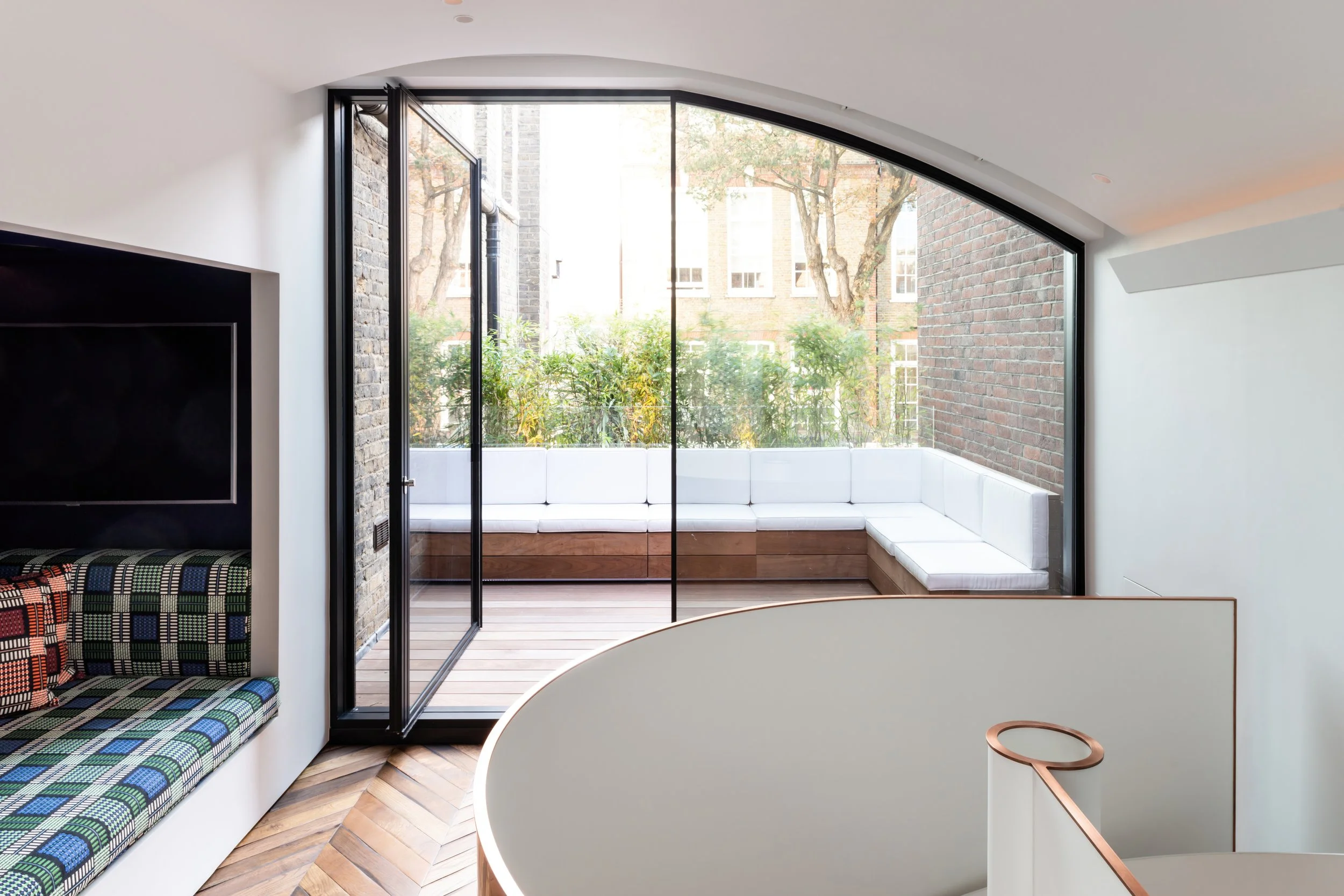 Sunlit interior with a large floor-to-ceiling glass door leading to a balcony with white cushioned seating, brick walls, and wooden flooring.