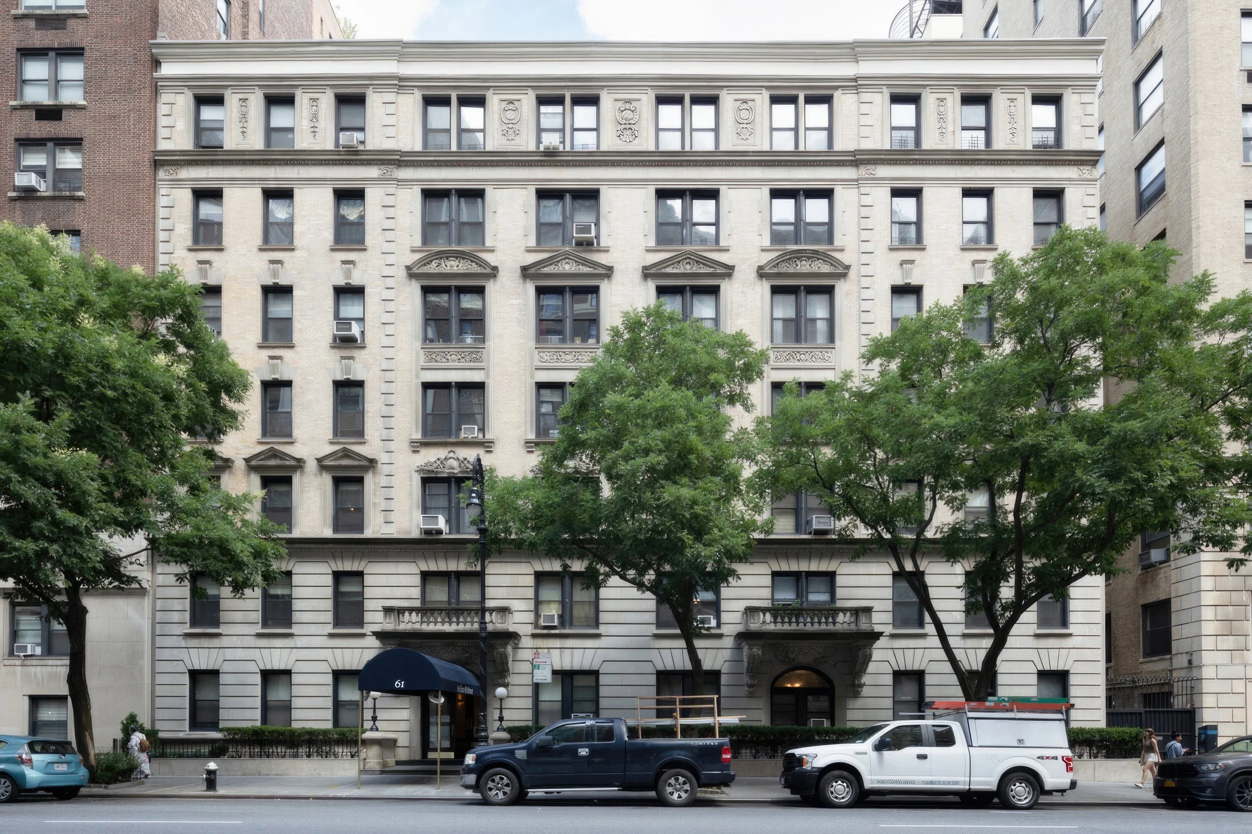 A historic multi-story residential building with decorated window frames, situated on a city street with trees, cars, and pedestrians.