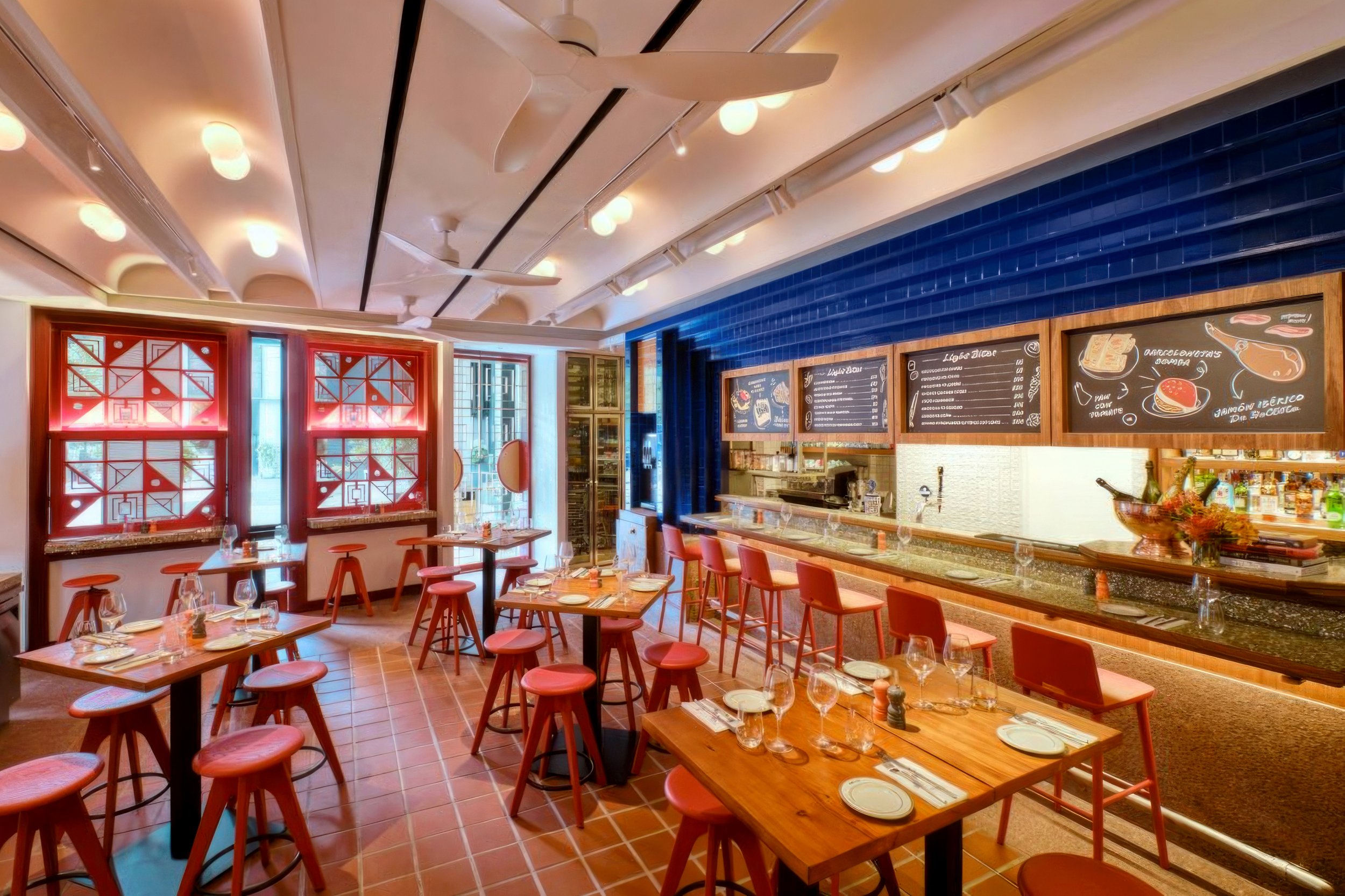 Empty restaurant with wooden tables and red stools, chalkboard menus on the wall, and decorative windows with red frames.