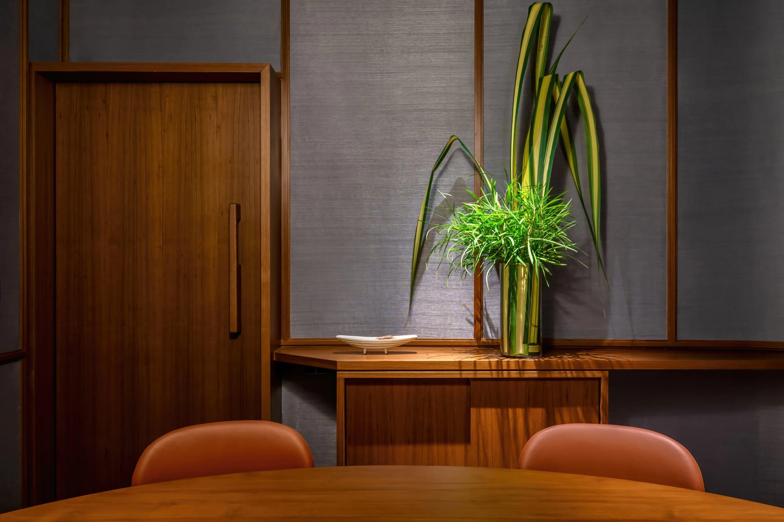Interior of a dining or conference room featuring a round wooden table, two orange chairs, a wooden cabinet, and a tall green potted plant with long leaves against a gray wall.