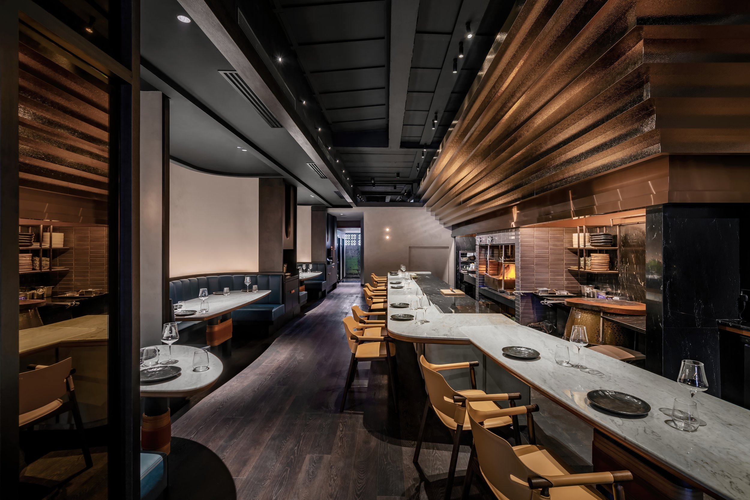 Interior of a modern restaurant with a marble high-top counter, wooden chairs, and a cooking area with a fireplace, dark wood floors, and contemporary design elements.