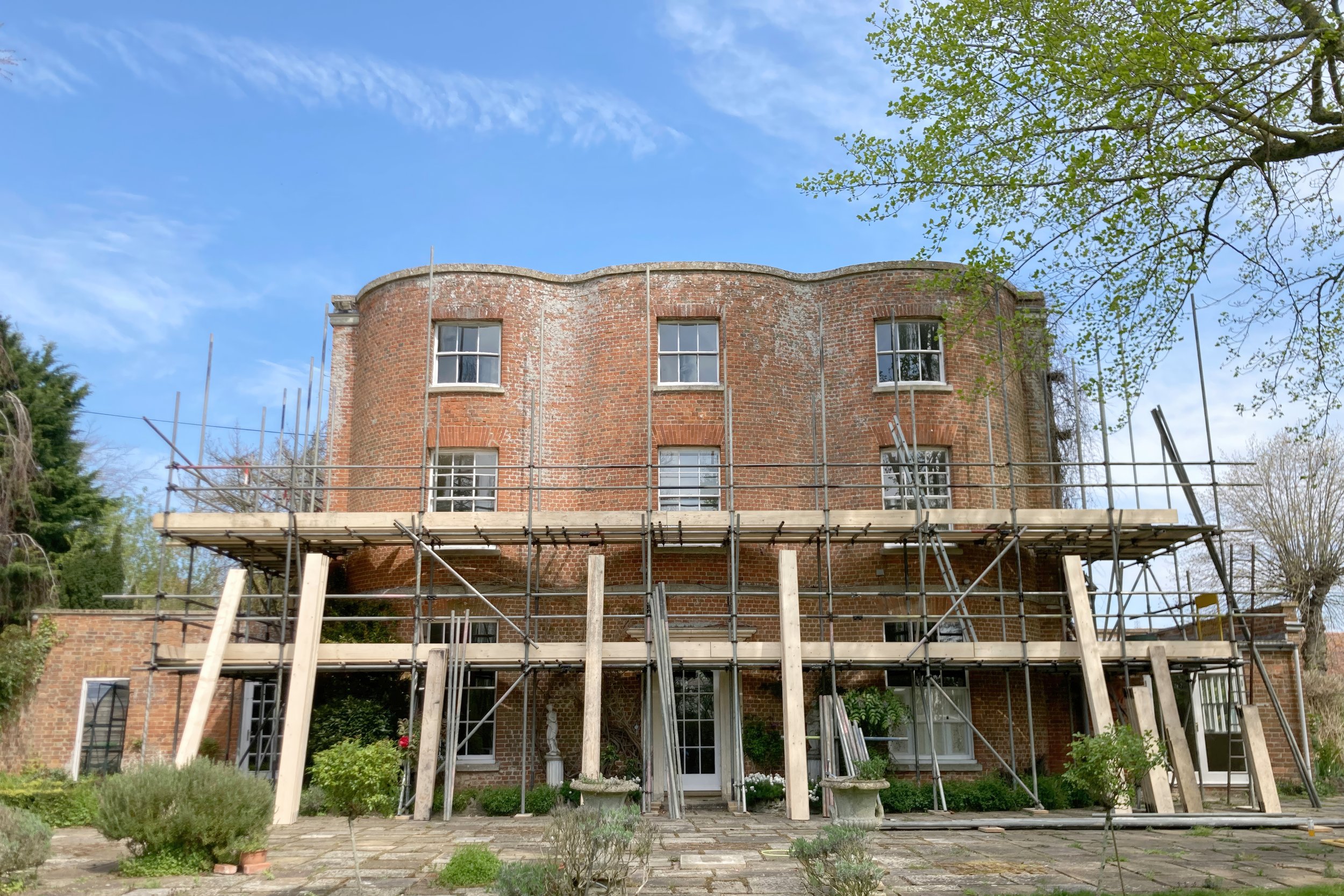 A brick building under renovation with scaffolding around it, set against a blue sky with some clouds and trees.