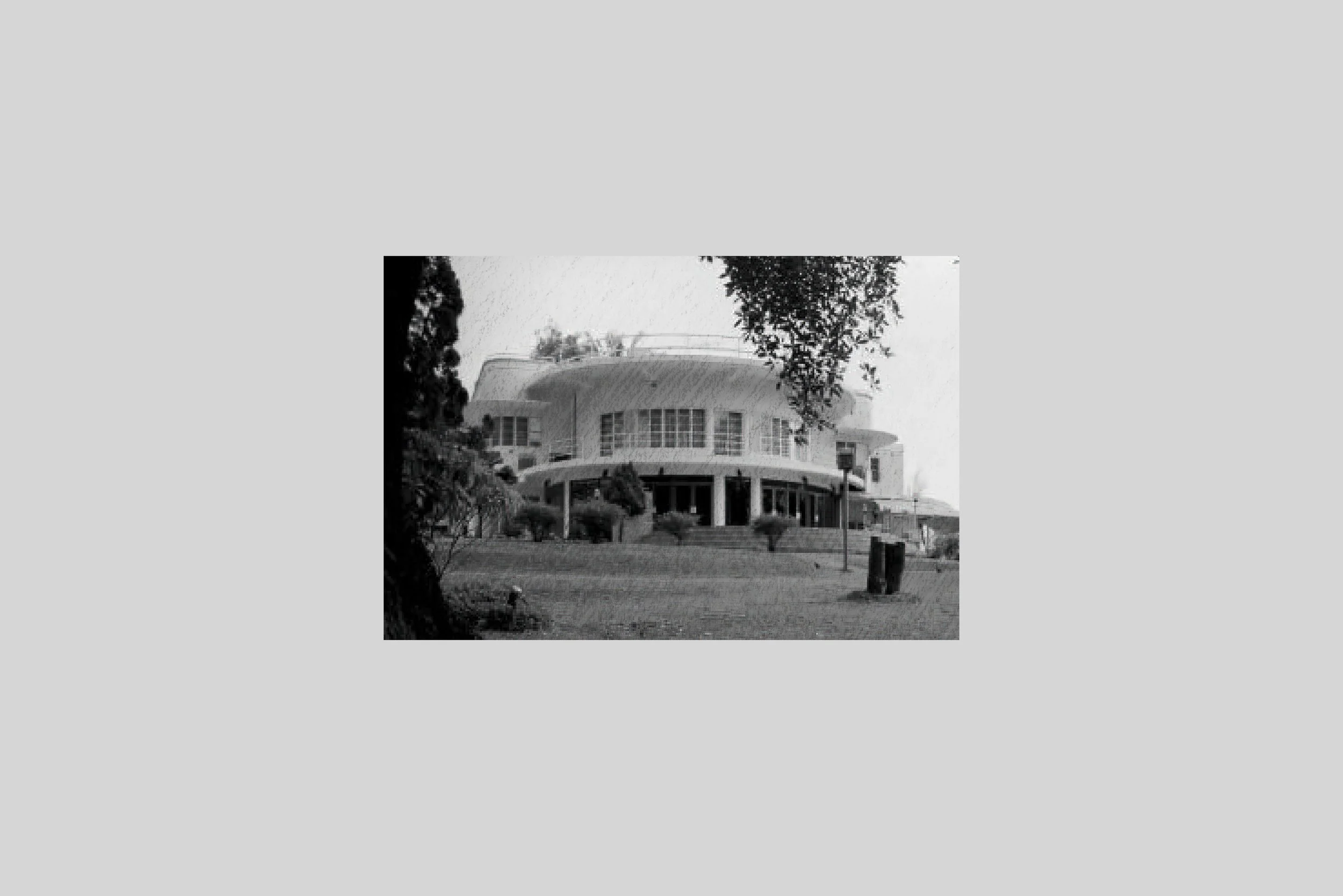 Black and white photo of a modern house with large windows and a curved roof, surrounded by trees and shrubs.