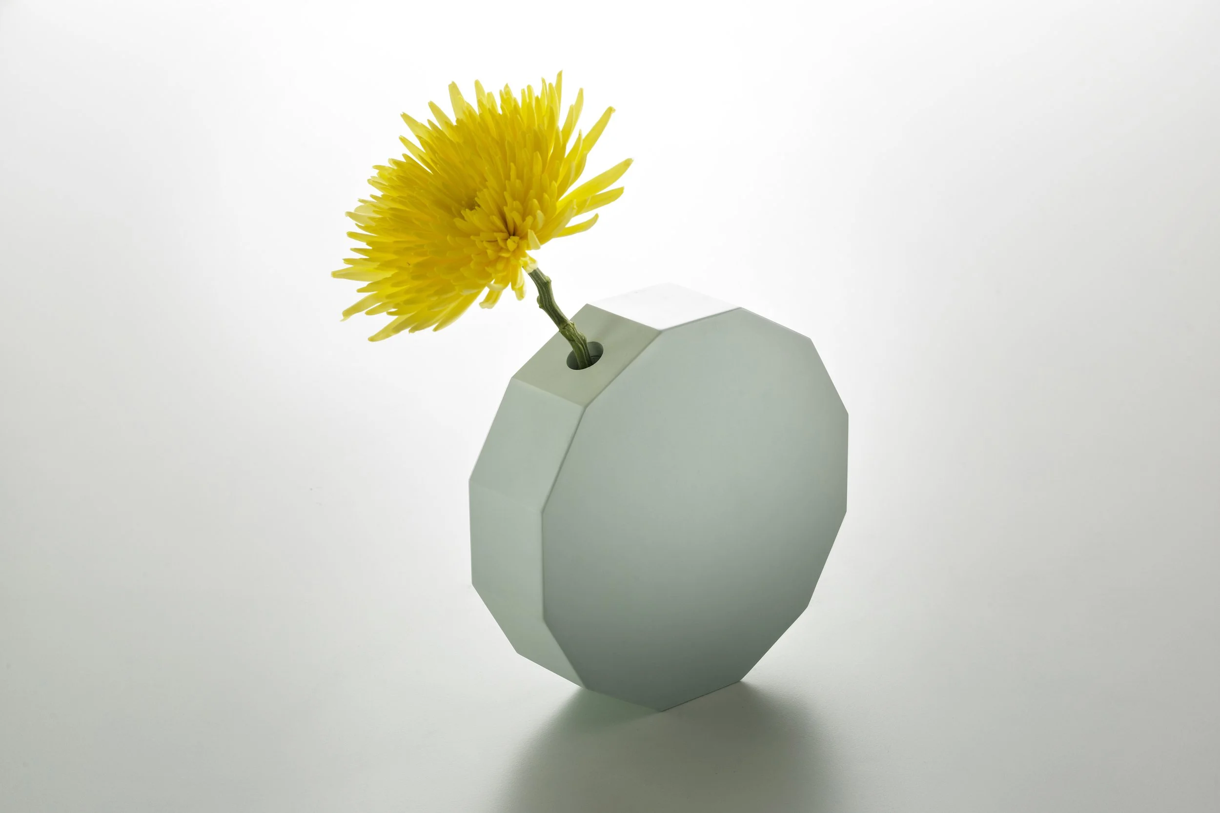 A geometric white vase holding a single yellow flower, set against a plain white background.