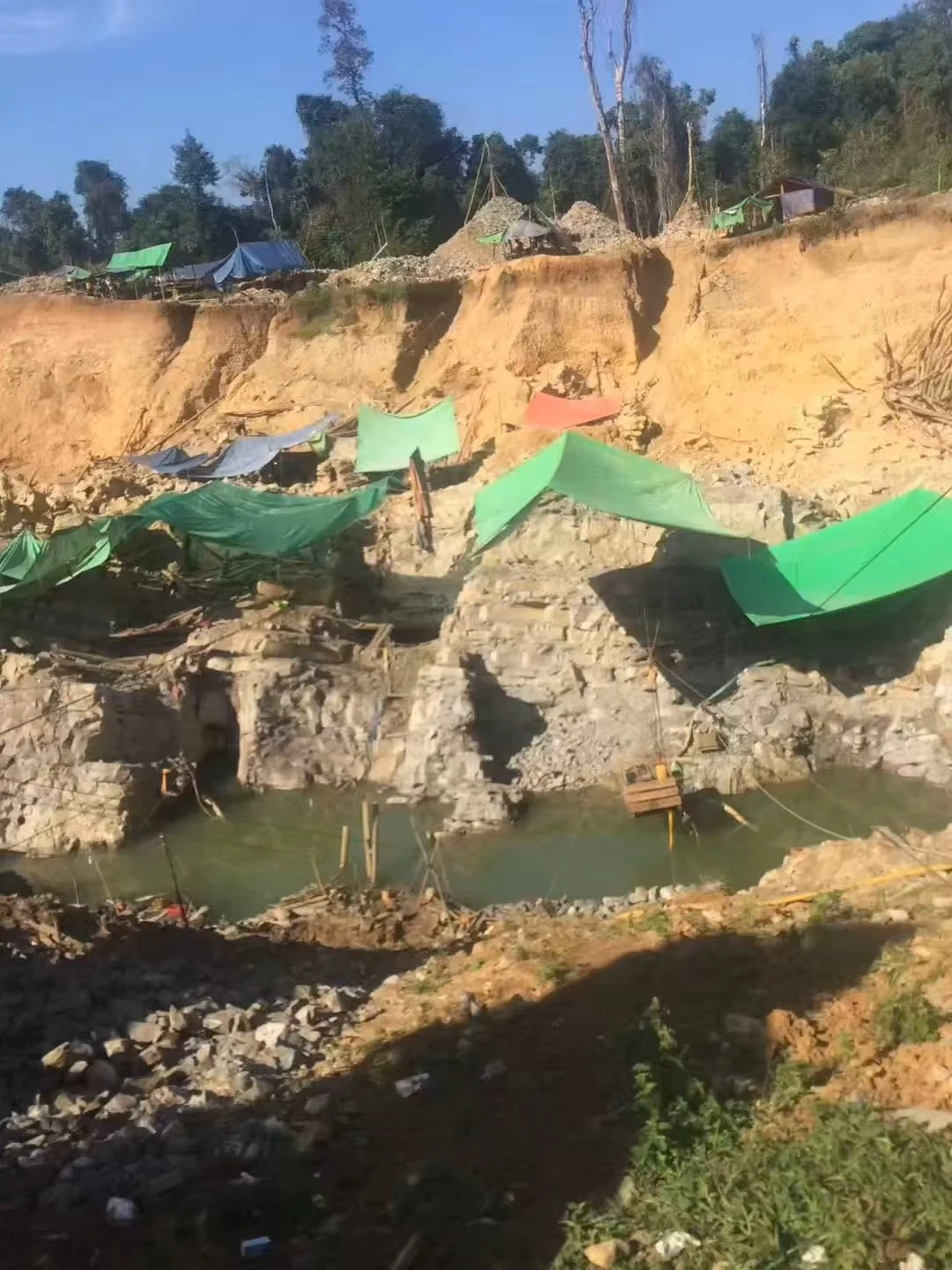 A partially collapsed hillside with green and blue tarps used as makeshift covers, and some wooden structures around a water-filled area, in a rural setting with trees in the background.