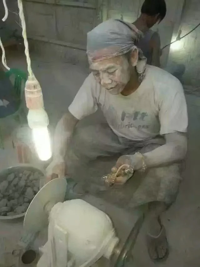 A person operating a traditional stone grinder, with a bowl of dried black berries nearby, in a dusty workshop setting.