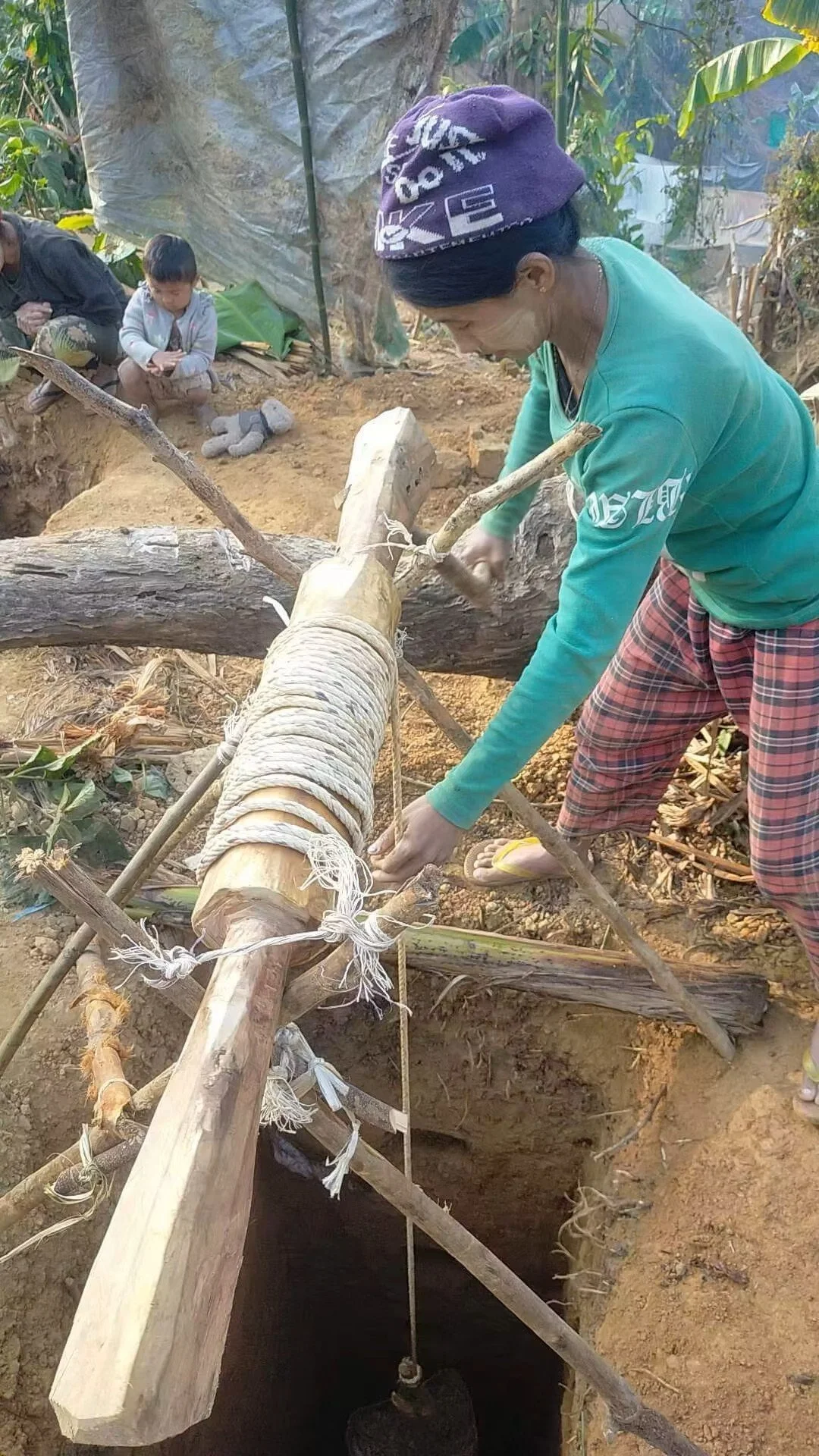 A woman in plaid pants and a turquoise shirt working on a wooden structure with ropes, possibly in a rural outdoor setting. Children are sitting nearby, some using a tablet, surrounded by plants and dirt.