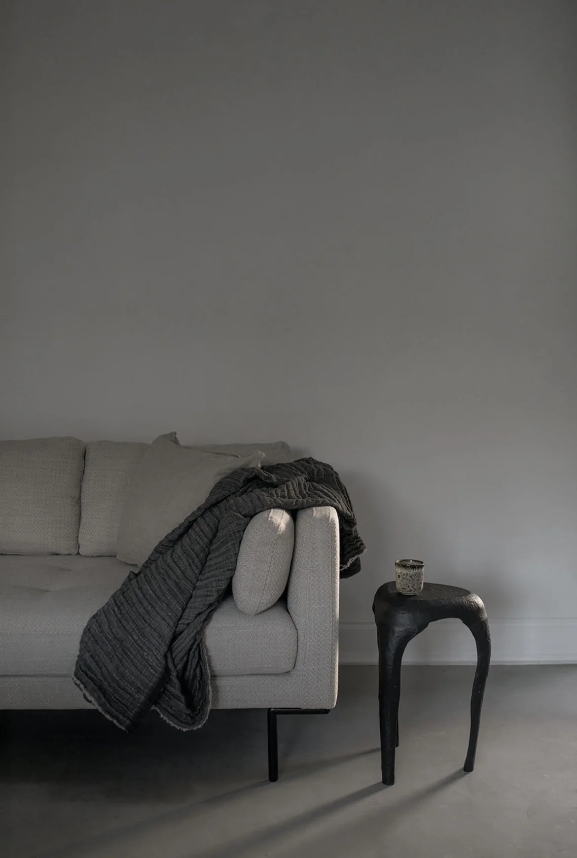 A light gray sofa with cushions and a folded dark gray blanket draped over one arm, a small black side table with a textured ceramic cup, all set against a plain gray wall.