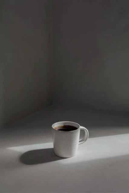 A white mug filled with black coffee on a light-colored surface, casting a shadow against a plain wall.