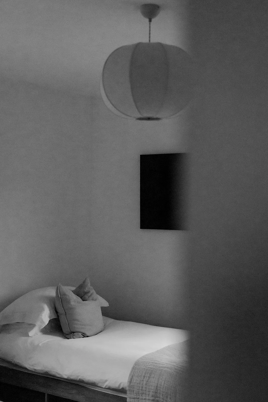 A bedroom with a bed and pillows, a wall-mounted flat screen TV, a ceiling light fixture, and a lamp, all in black and white.