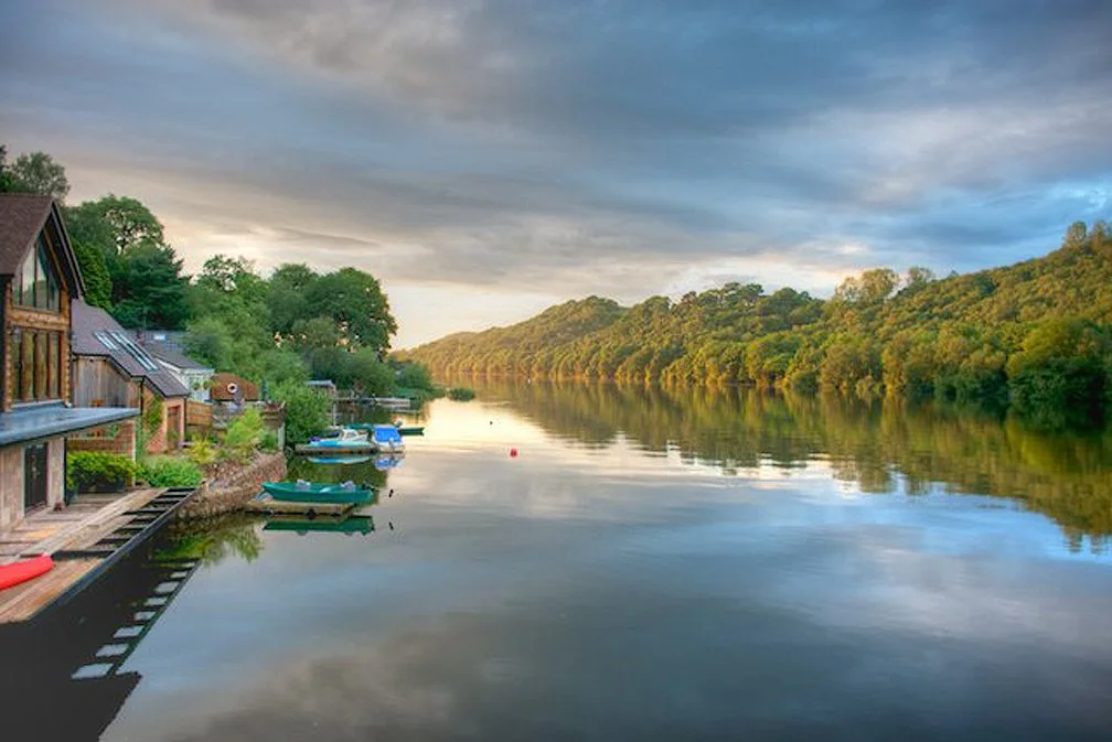 A serene river scene during dusk with houses on the left and trees on the right, calm water reflecting the cloudy sky.