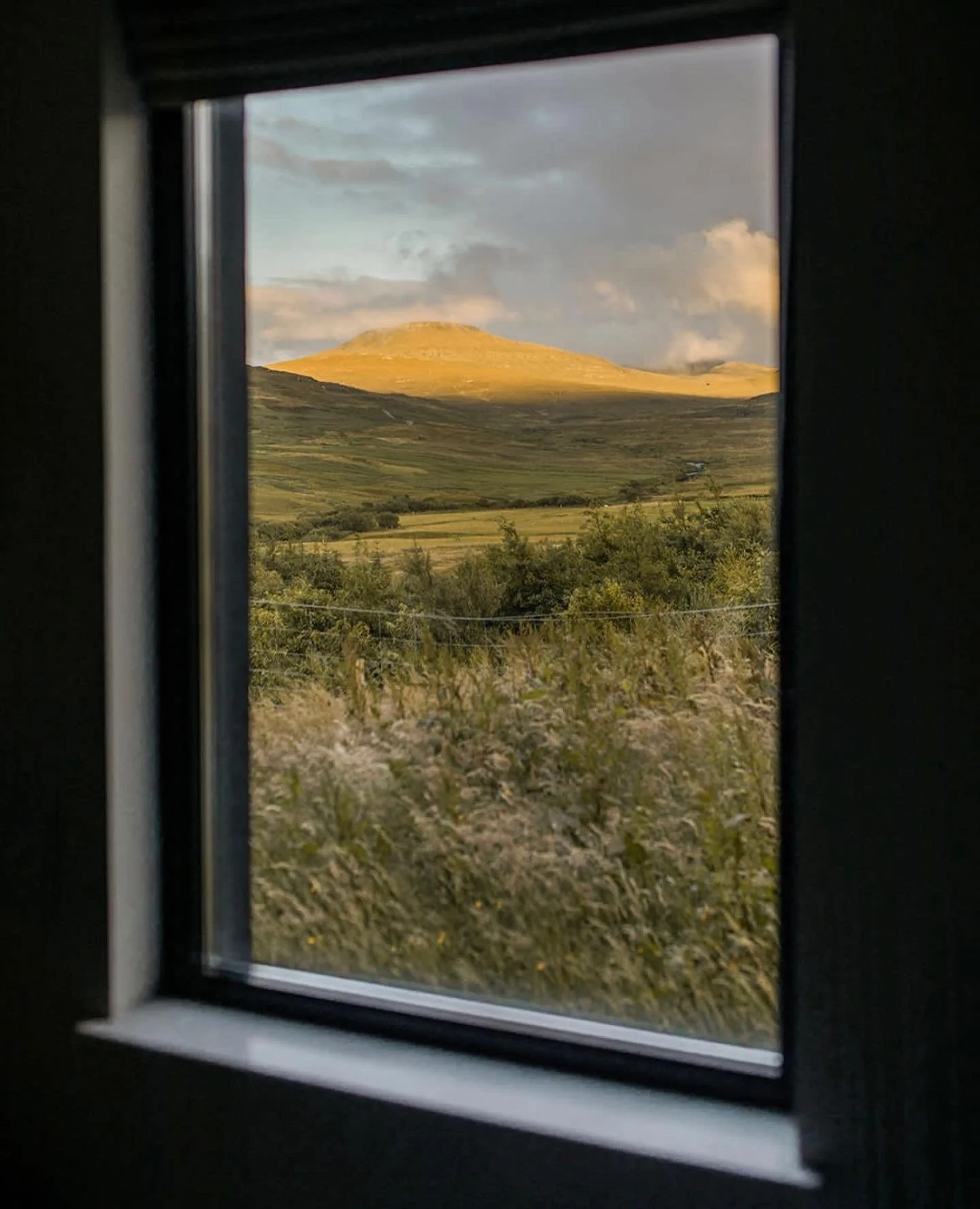 Scenic view of rolling hills, some with grass and others with shrubs, seen through a window with black frame.
