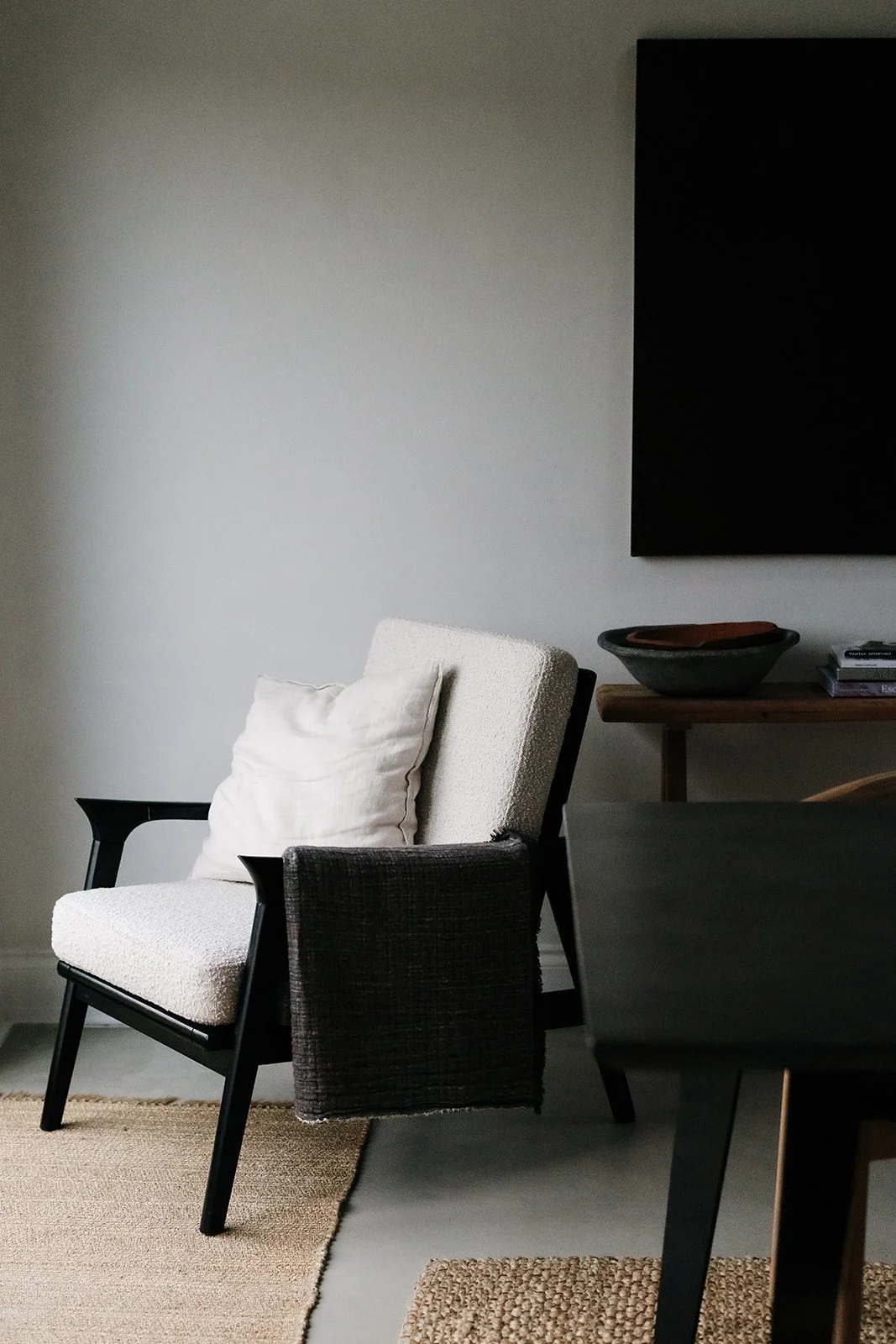 A modern living room corner featuring a black and white armchair with a cream cushion, a woven black side table, a wooden console with a large black bowl, and a stack of books. A wall-mounted TV is visible on a plain gray wall.