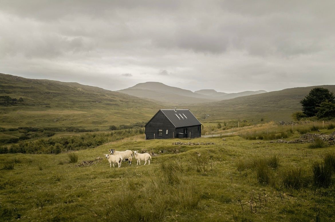 A black house on a grassy landscape with a few sheep and mountains in the background under a cloudy sky.