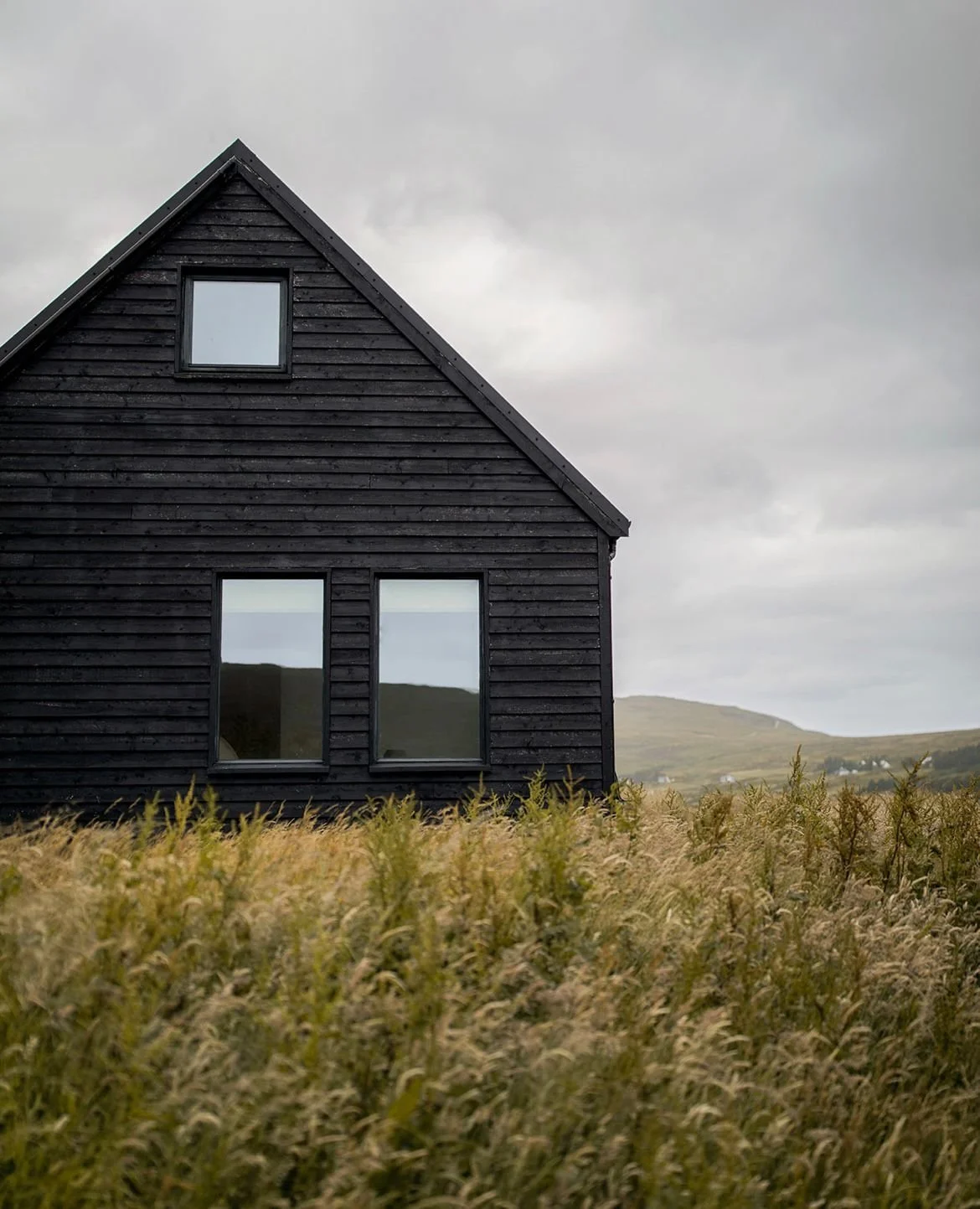 A black wooden house with three windows, situated in an open field with tall grass, under a cloudy sky, with hills in the background.