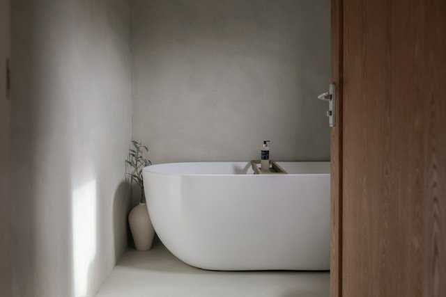 A modern, white freestanding bathtub with a faucet, next to a potted plant, in a minimalist bathroom with concrete walls and a wooden door.