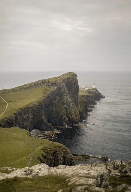 Cliffs along a coastline with a lighthouse in the distance, under a cloudy sky.