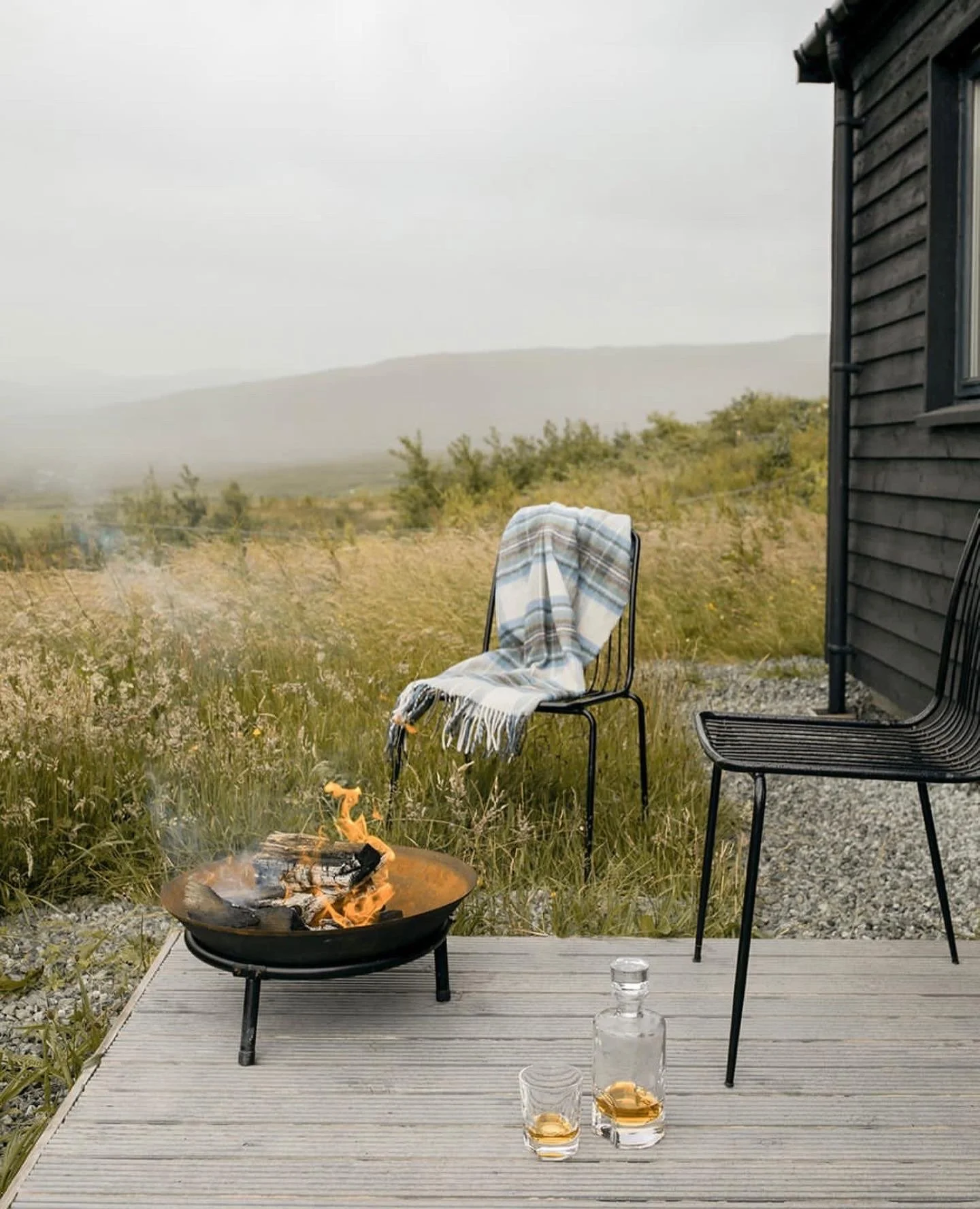 A small outdoor patio with two black chairs, one draped with a striped blanket. There is a portable fire pit with a small fire. Two glasses of alcohol are on a wooden deck, near a black house with horizontal siding. The background shows a foggy landscape with shrubs and a distant mountain.