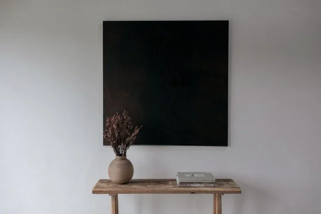 An empty black square canvas on a white wall with a wooden table below holding a vase with dried flowers and a closed book or album.