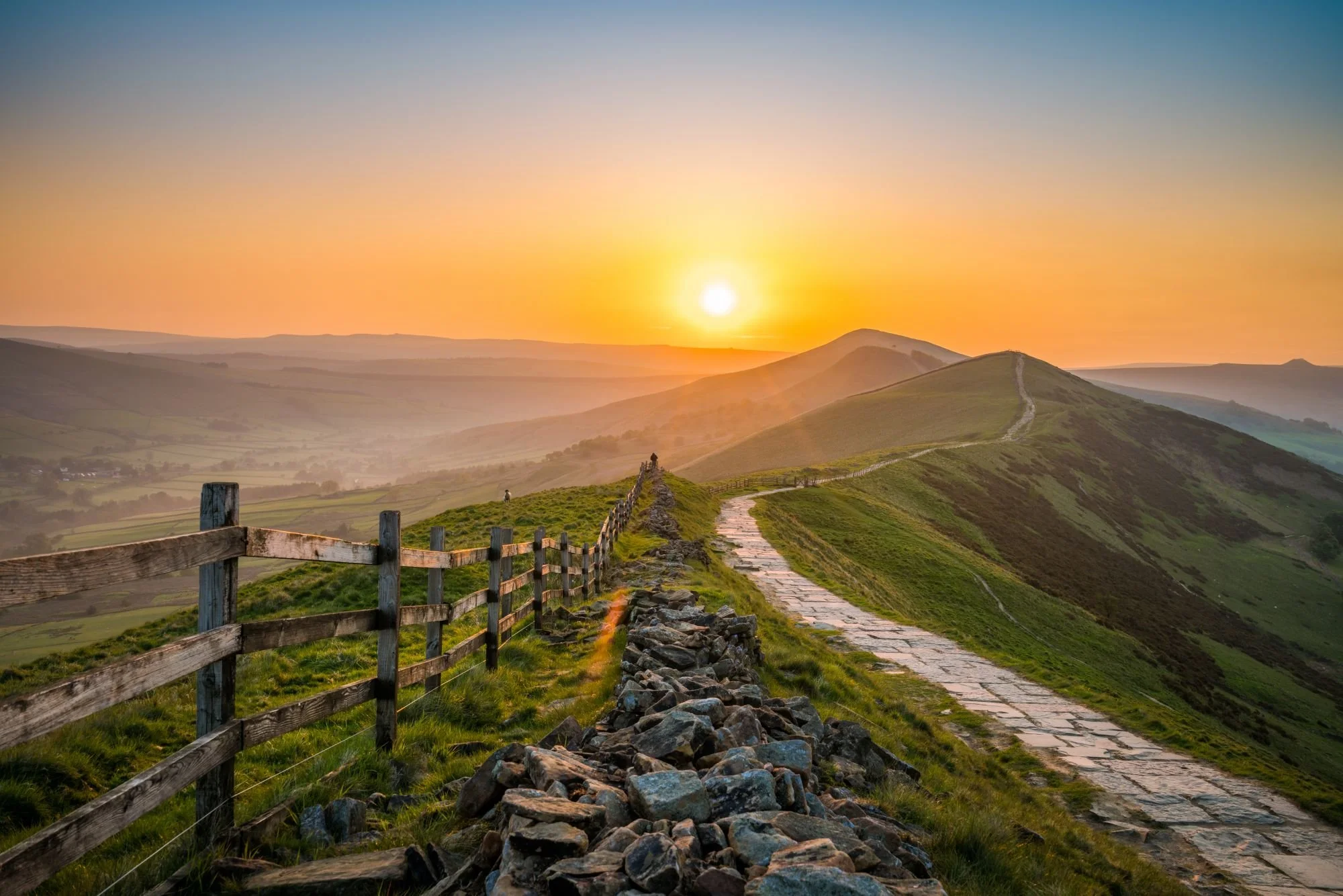 Sunset over rolling green hills with a stone-paved country path and wooden fence in the foreground.