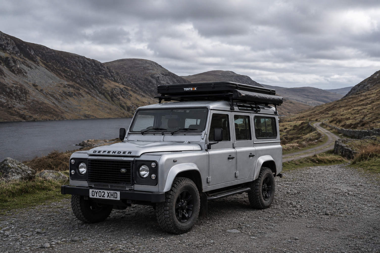 Silver Land Rover Defender parked on a gravel road beside a lake with mountains in the background, overcast sky.