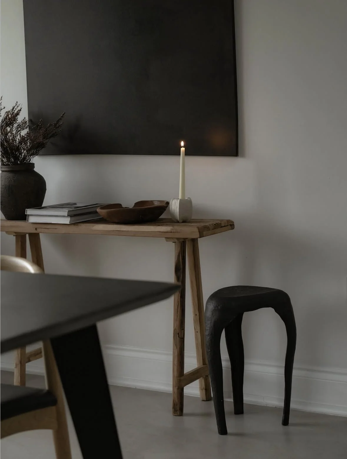 A minimalist interior scene with a wooden table holding a large black vase with dried flowers, a stack of books, a wooden bowl, and a lit candle in a textured white holder. A black framed chalkboard is on the wall behind, with a black wooden chair in front of the table.
