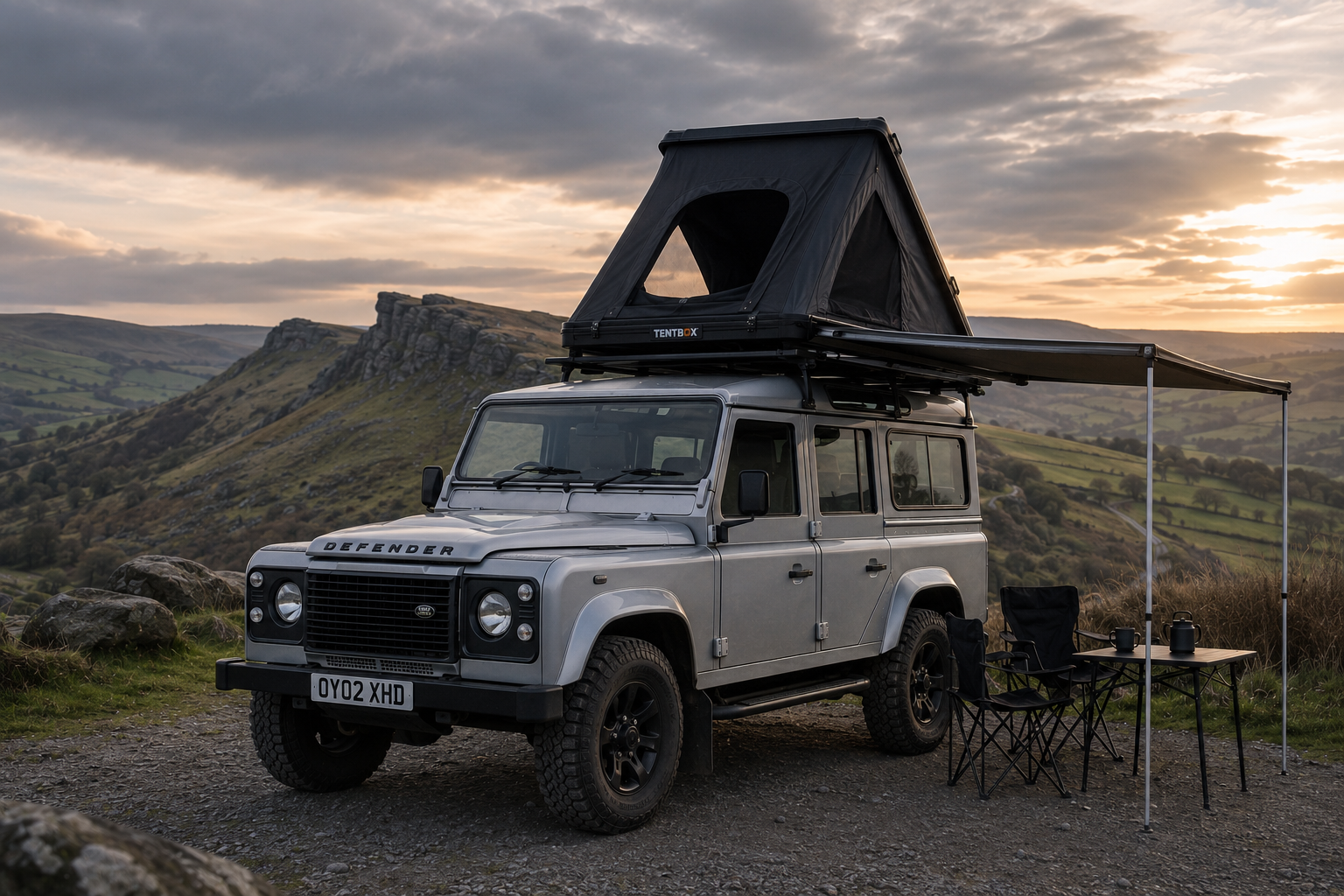 A silver Land Rover Defender with a rooftop tent and an awning set up in a scenic outdoor landscape with hills and rocks at sunset, with a small table and chairs outside.