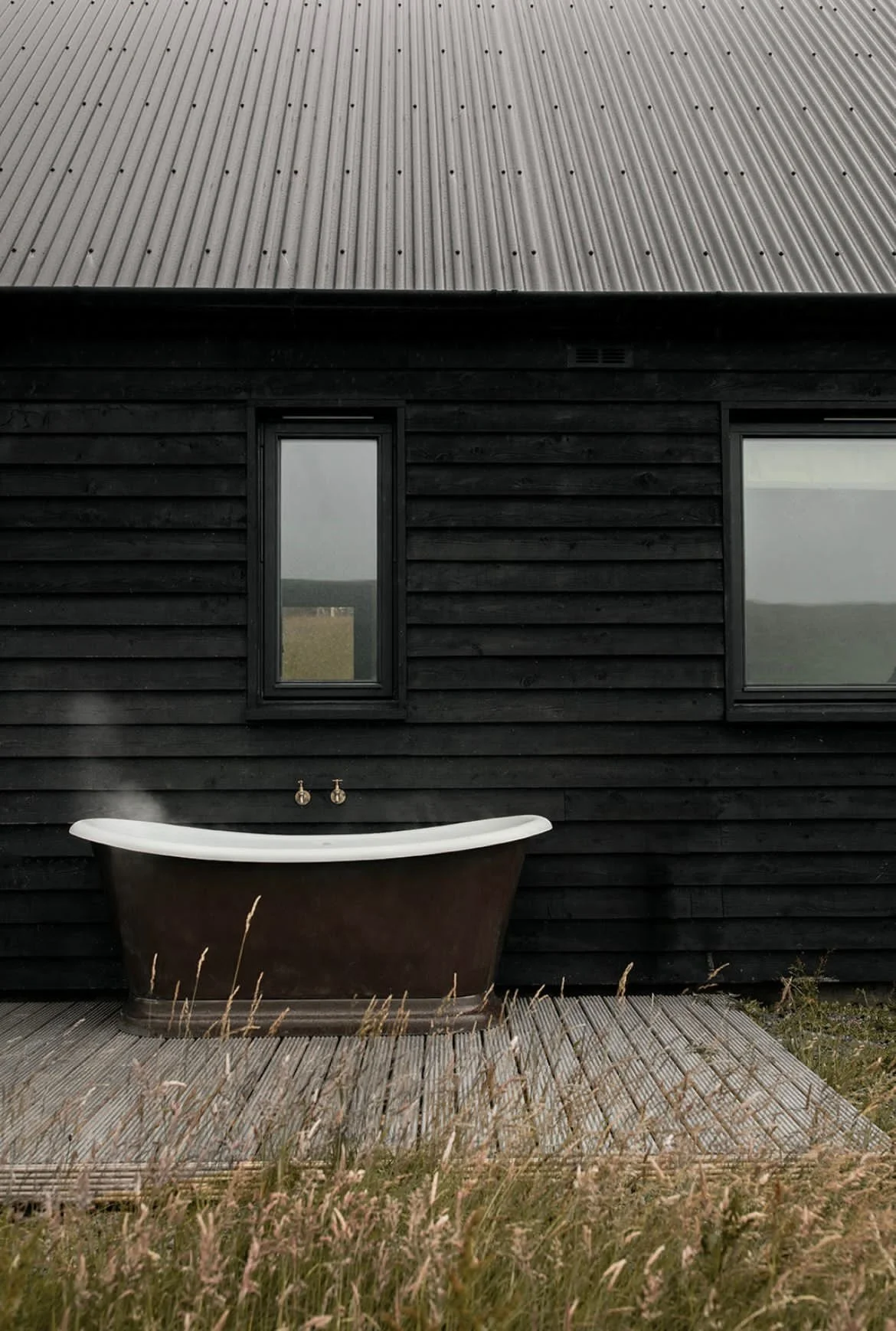 Exterior of a black wooden house with a silver metal roof, featuring two small rectangular windows and an outdoor bathtub on a wooden deck, with tall grasses in the foreground under a cloudy sky.