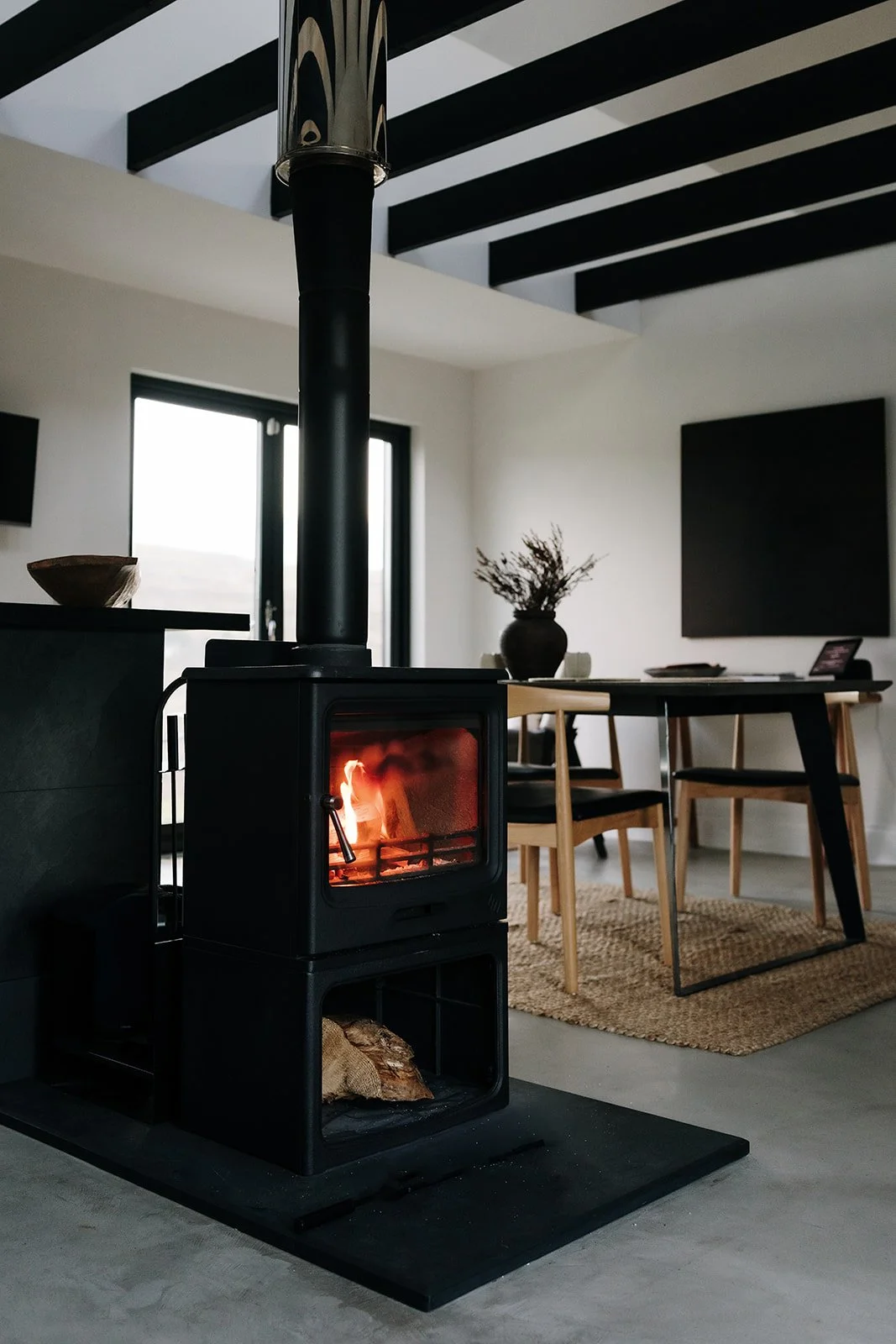 A modern living room with a black wood stove burning, a dining area with a wooden table, black and beige chairs, a large black television on the wall, and a vase with dried flowers on the table. Natural light coming through windows with black frames.