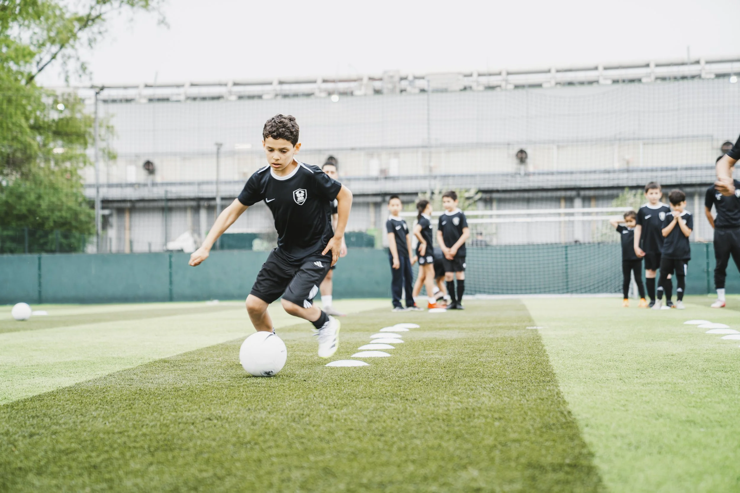 Young boy in black sports attire kicking a soccer ball on a grassy field during a practice or game, with other children and adults in the background.