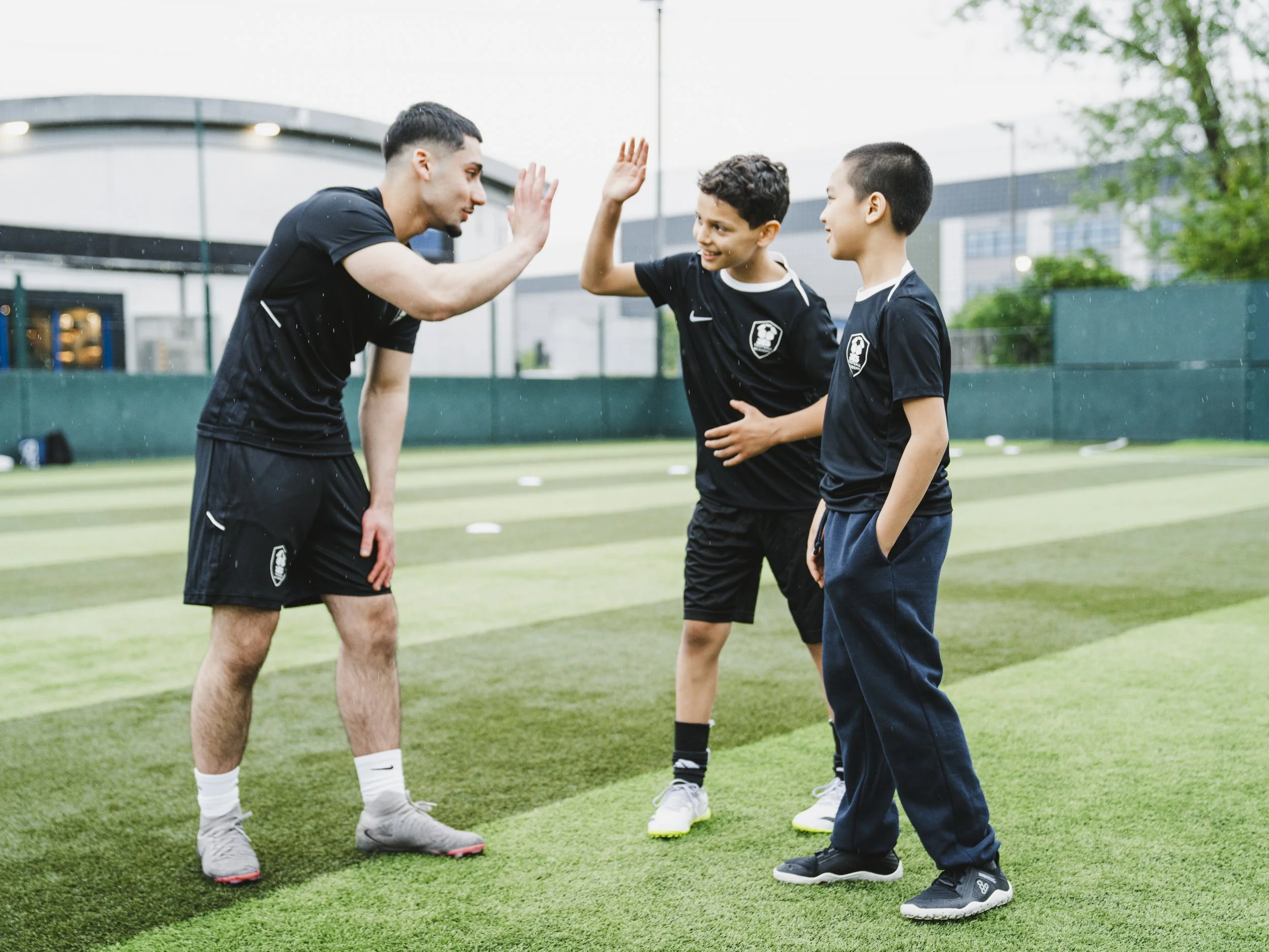 Two soccer players, one adult and one child, giving each other a high five on a soccer field with a modern building and trees in the background.