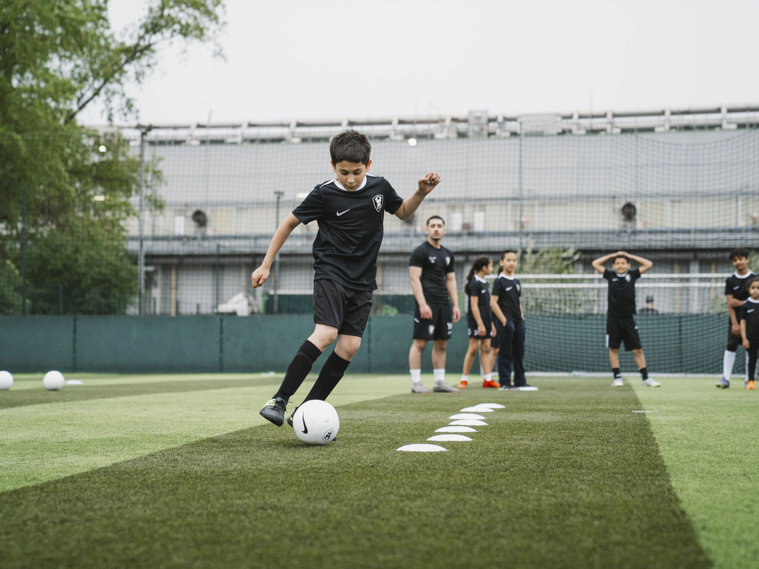 Young boy practicing soccer skills on the field, kicking a soccer ball with a line of small cones marking a training drill, with a group of children and a coach in the background.