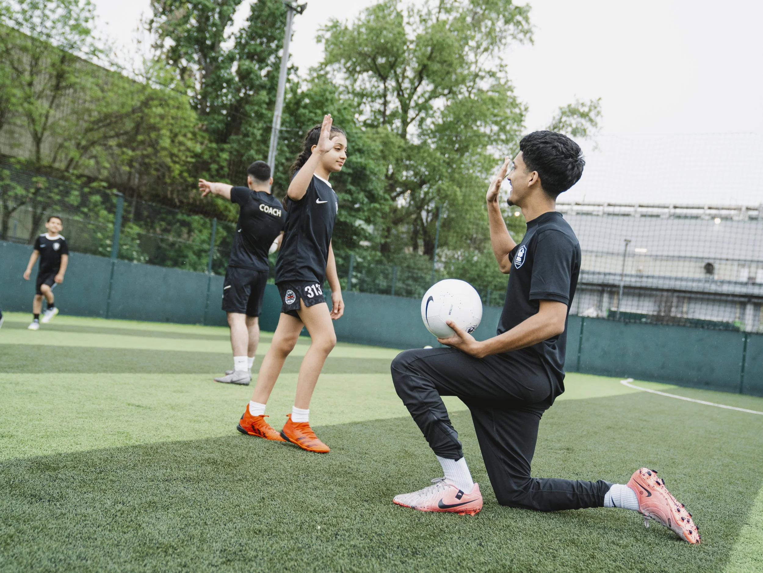 A soccer coach kneeling on one knee and giving a high five to a female soccer player on a green turf field, with two other children in the background, surrounded by trees and a fence.