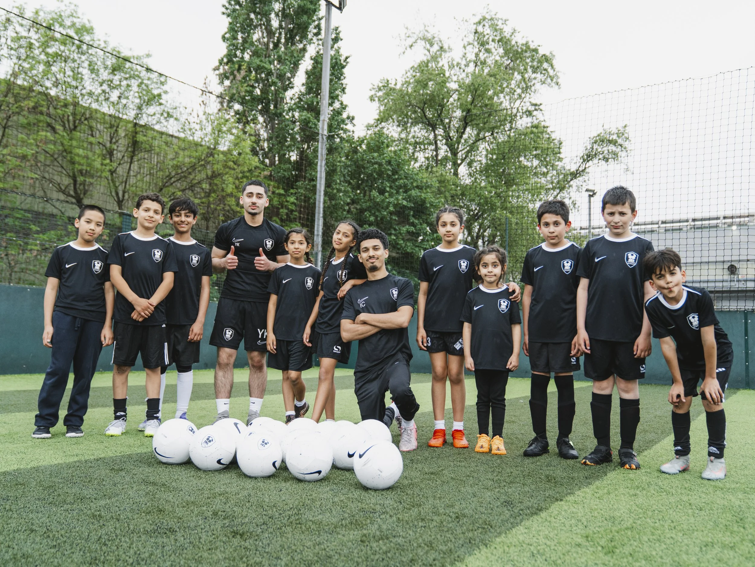 A youth soccer team with two coaches standing on a soccer field surrounded by trees, with soccer balls in front and a netted fence behind.