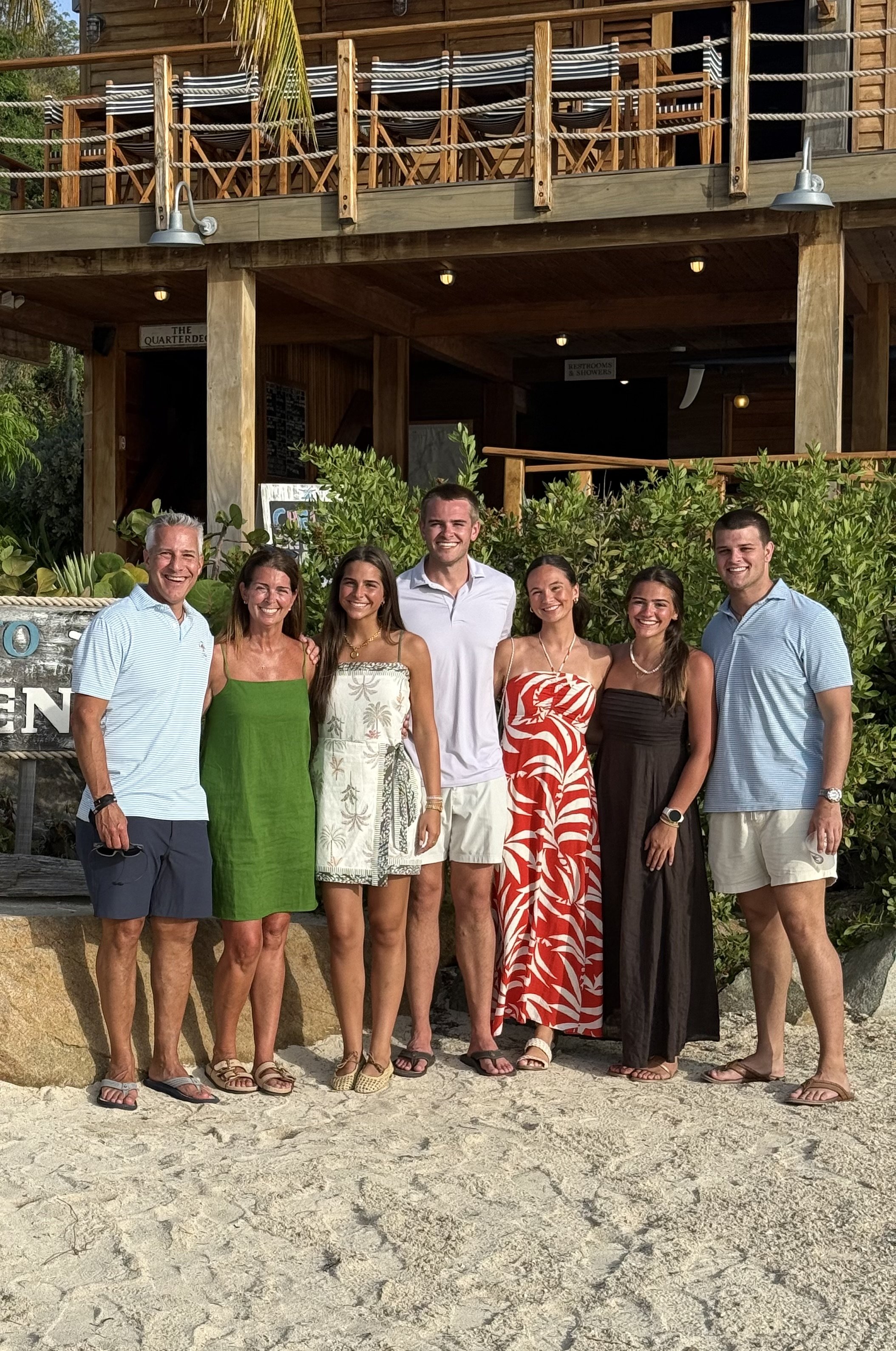 Group of seven people, three men and four women, standing on a sandy beach in front of a wooden building with a balcony and greenery. They are smiling and dressed in summer attire.