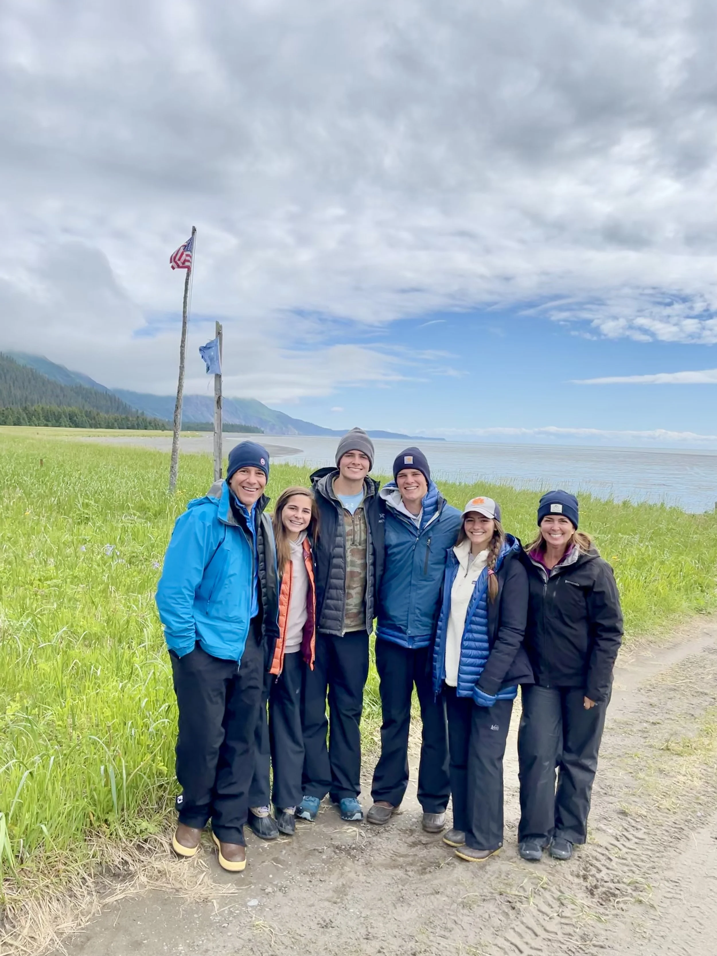 A group of seven people standing outdoors on a grassy area near a body of water, dressed in outdoor clothing and smiling at the camera, with mountains and cloudy sky in the background.
