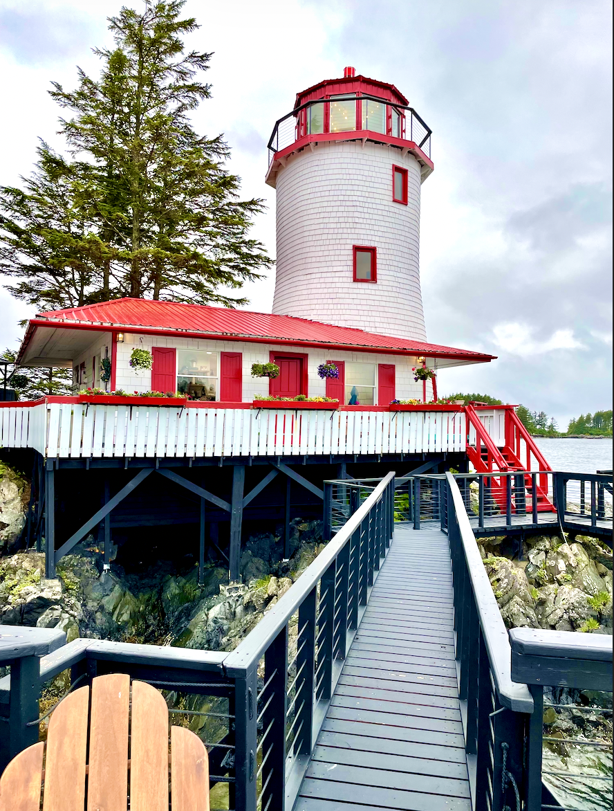 A lighthouse with a white body and red roof, surrounded by a white picket fence with red trim, accessed via a walkway with black railings.