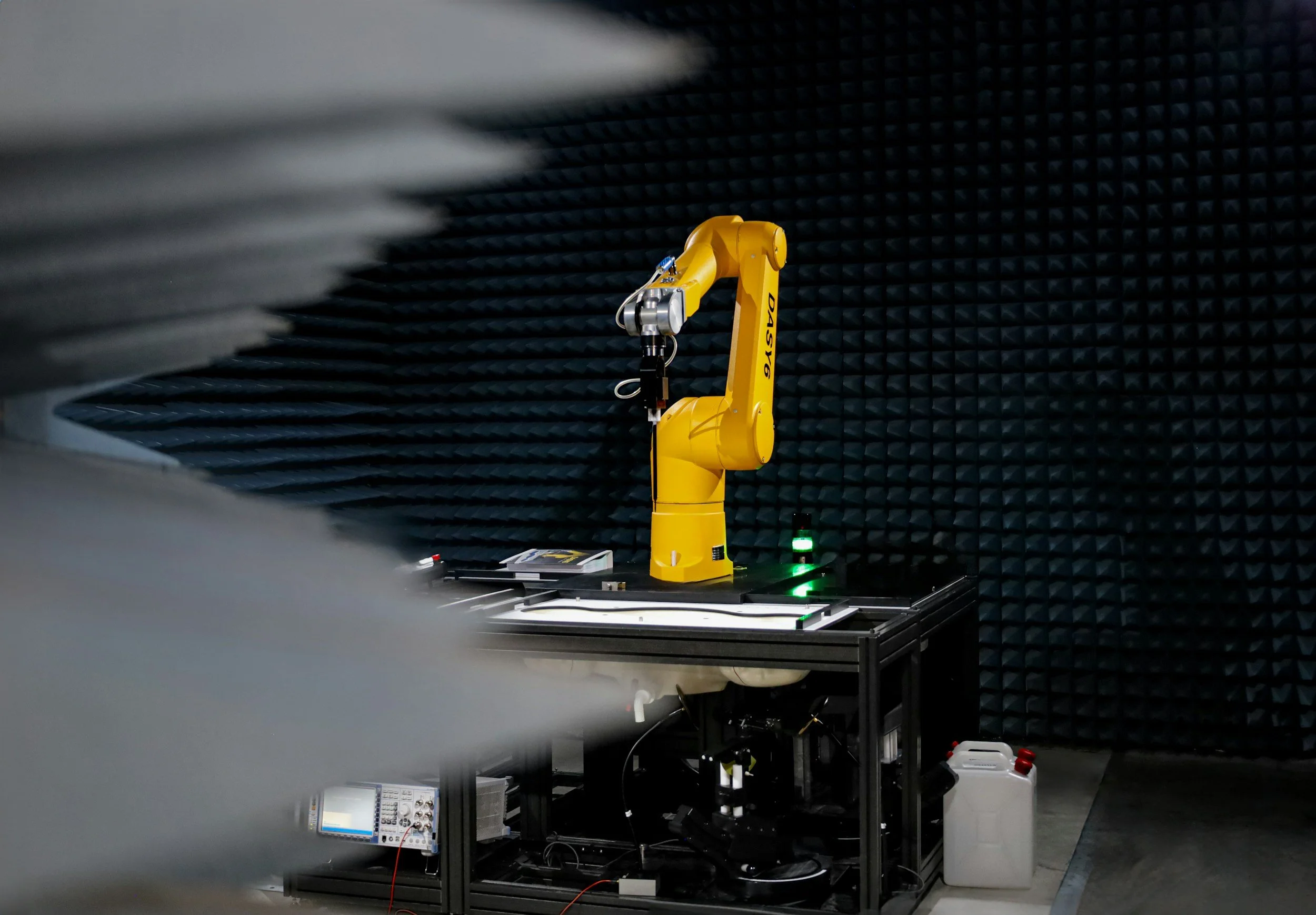 Yellow robotic arm inside an anechoic chamber with foam acoustic panels on the walls, mounted on a black metal table with electronic equipment and a white container nearby.