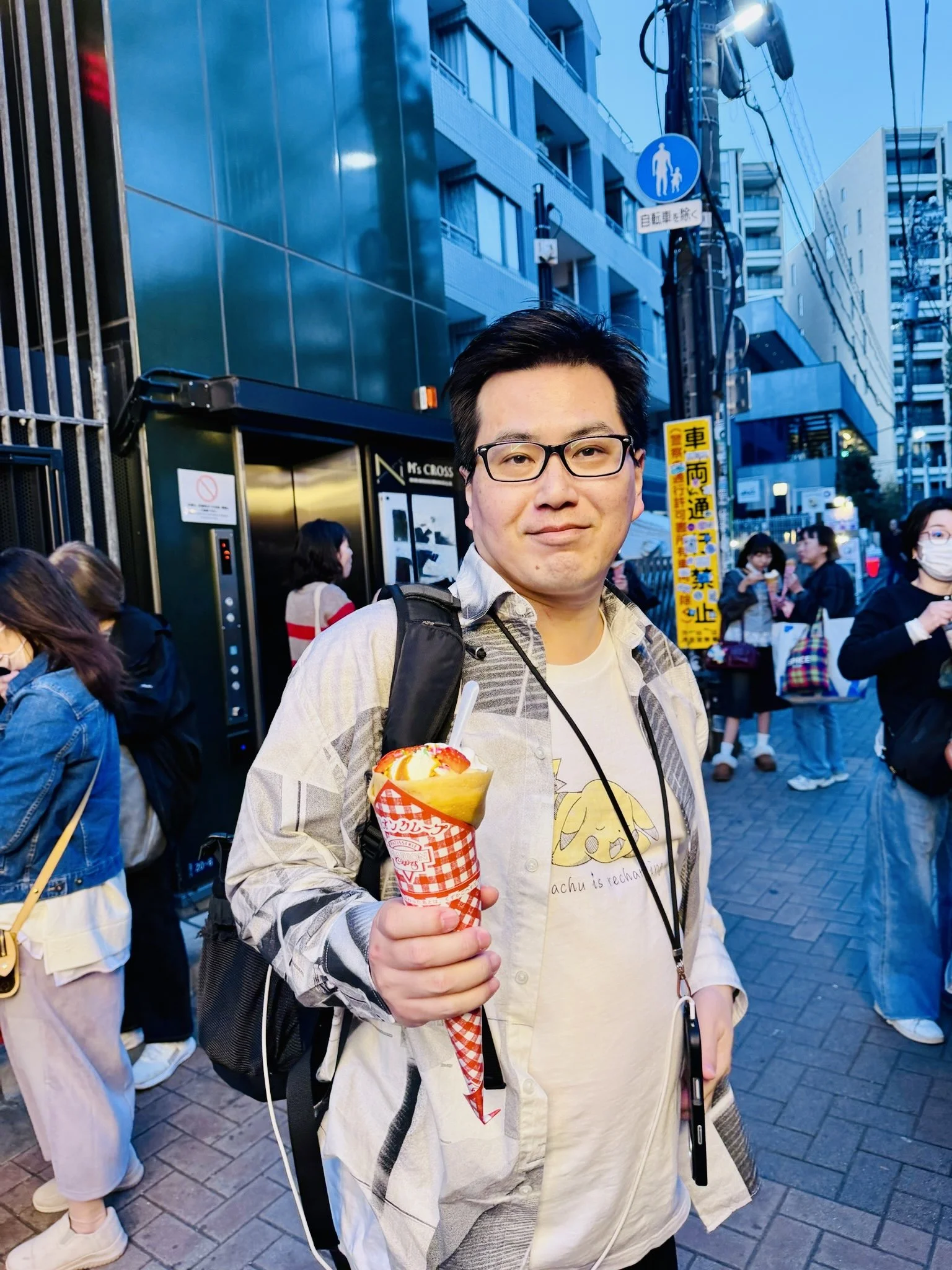 A Japanese man holding a strawberry and vanilla crape while standing on a busy city street in Harajuku, Tokyo, with other people in the background.