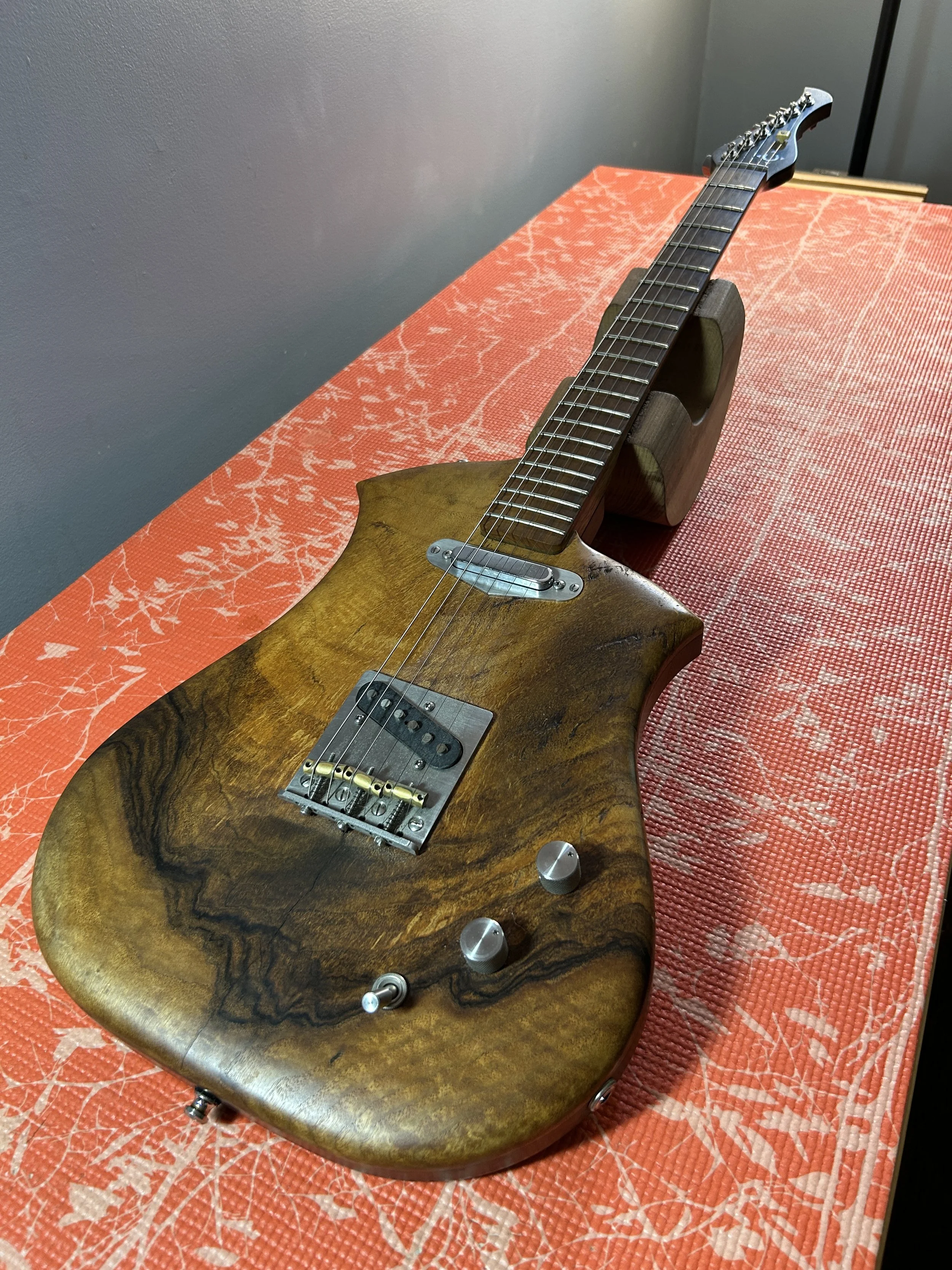 A wooden electric guitar resting on a wooden stand on an orange tablecloth with a leaf pattern, against a gray wall background.