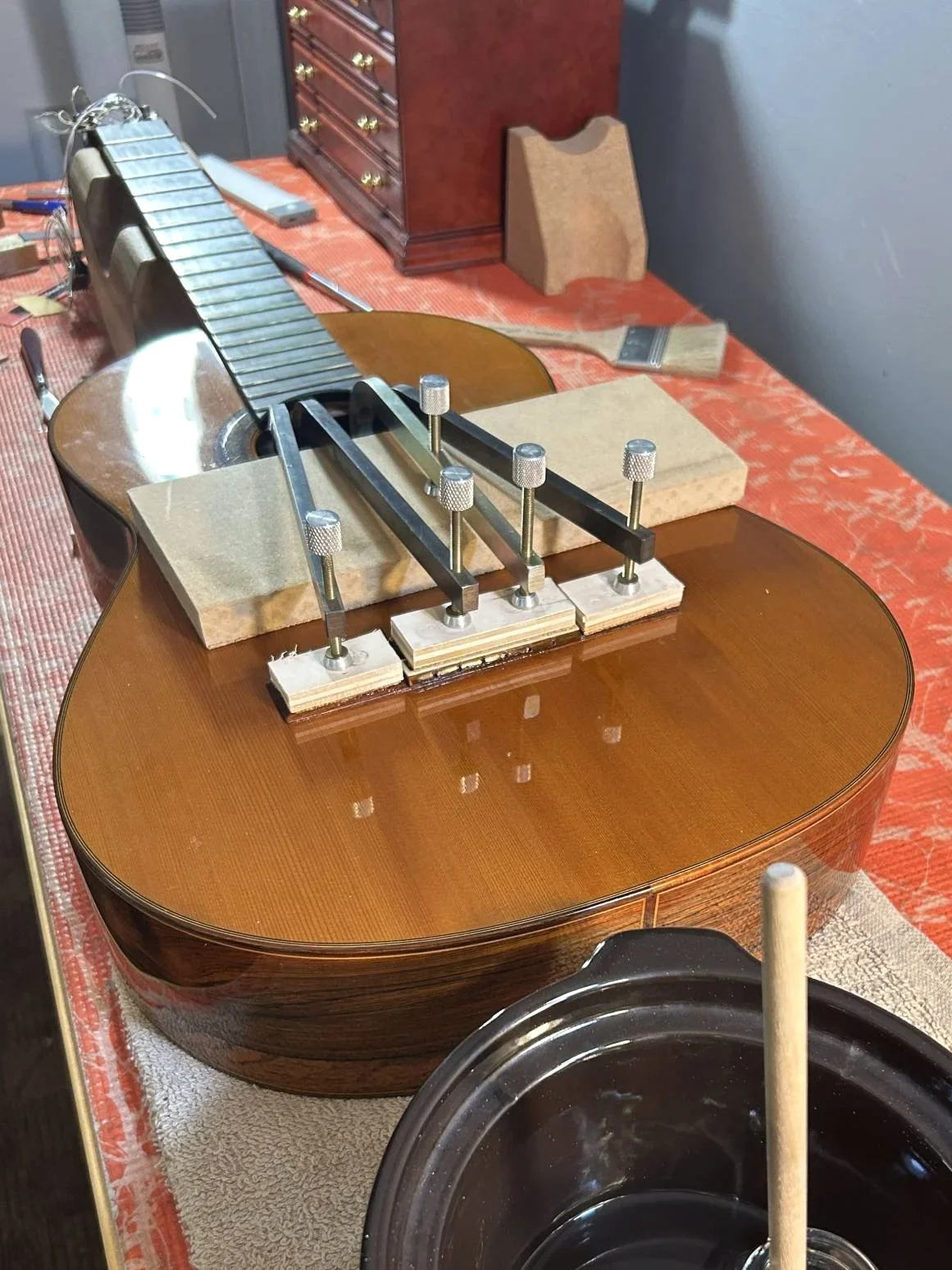 A guitar in the process of being glued or repaired, with clamps holding the body and neck together. Various tools and materials are on the work surface, including bricks and a brush.