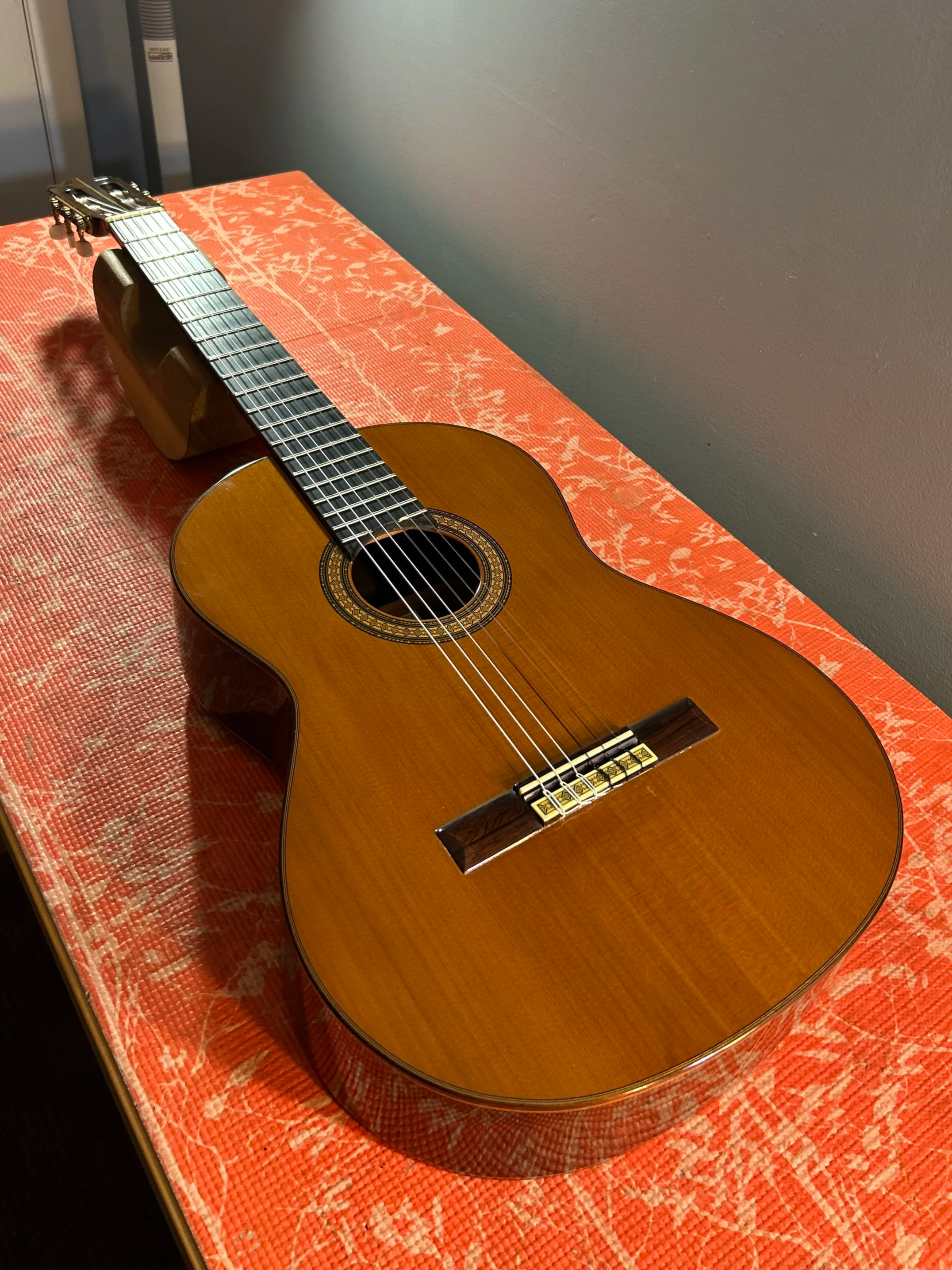 A classical acoustic guitar lying on an orange patterned pad against a gray wall.