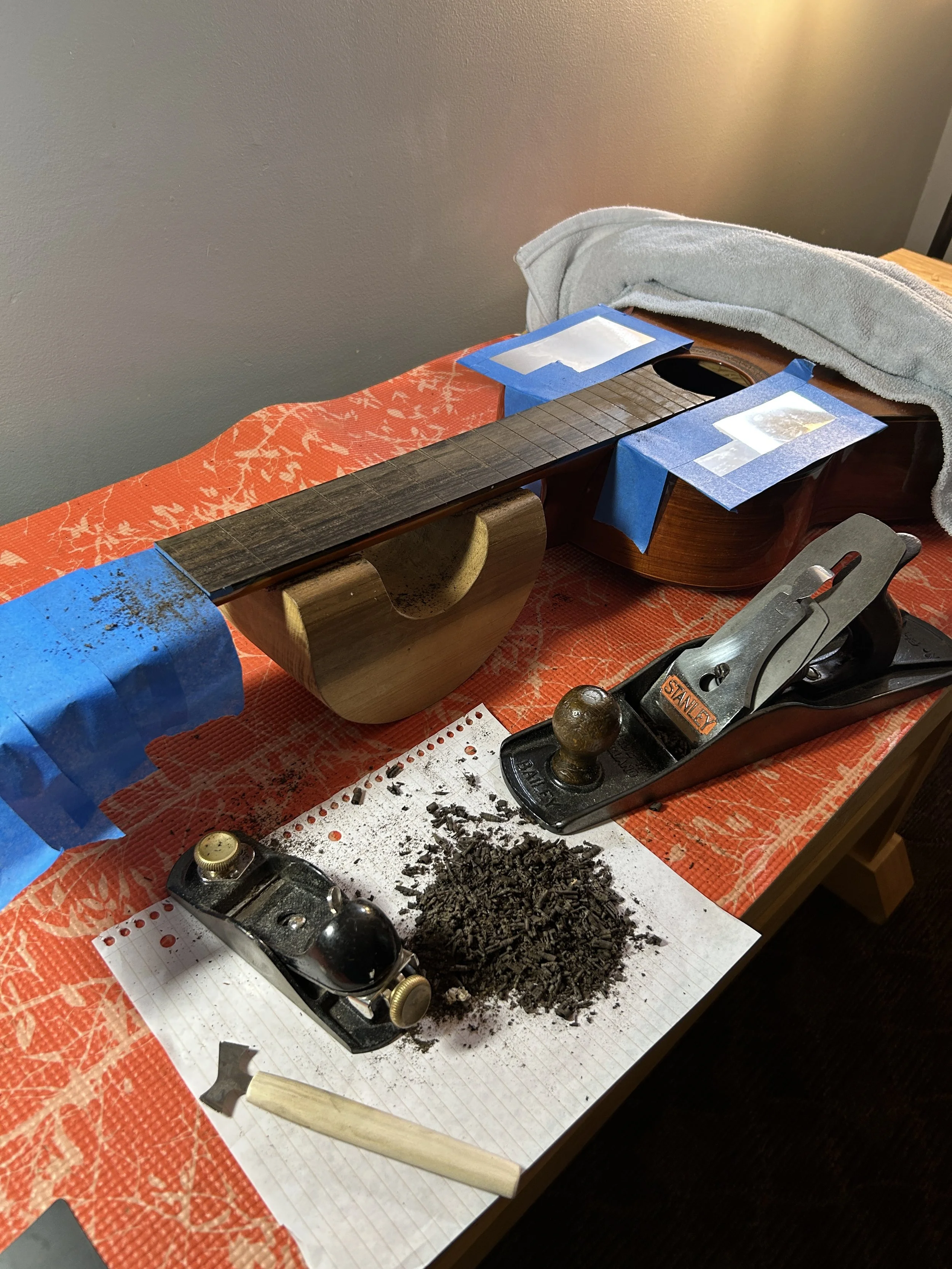 An acoustic guitar being sanded, with sanding tools, a pile of wood shavings, and masking tape on the guitar and workspace.