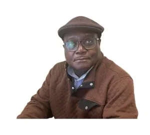 A man wearing glasses, a brown jacket, and a brown cap, sitting against a white background.
