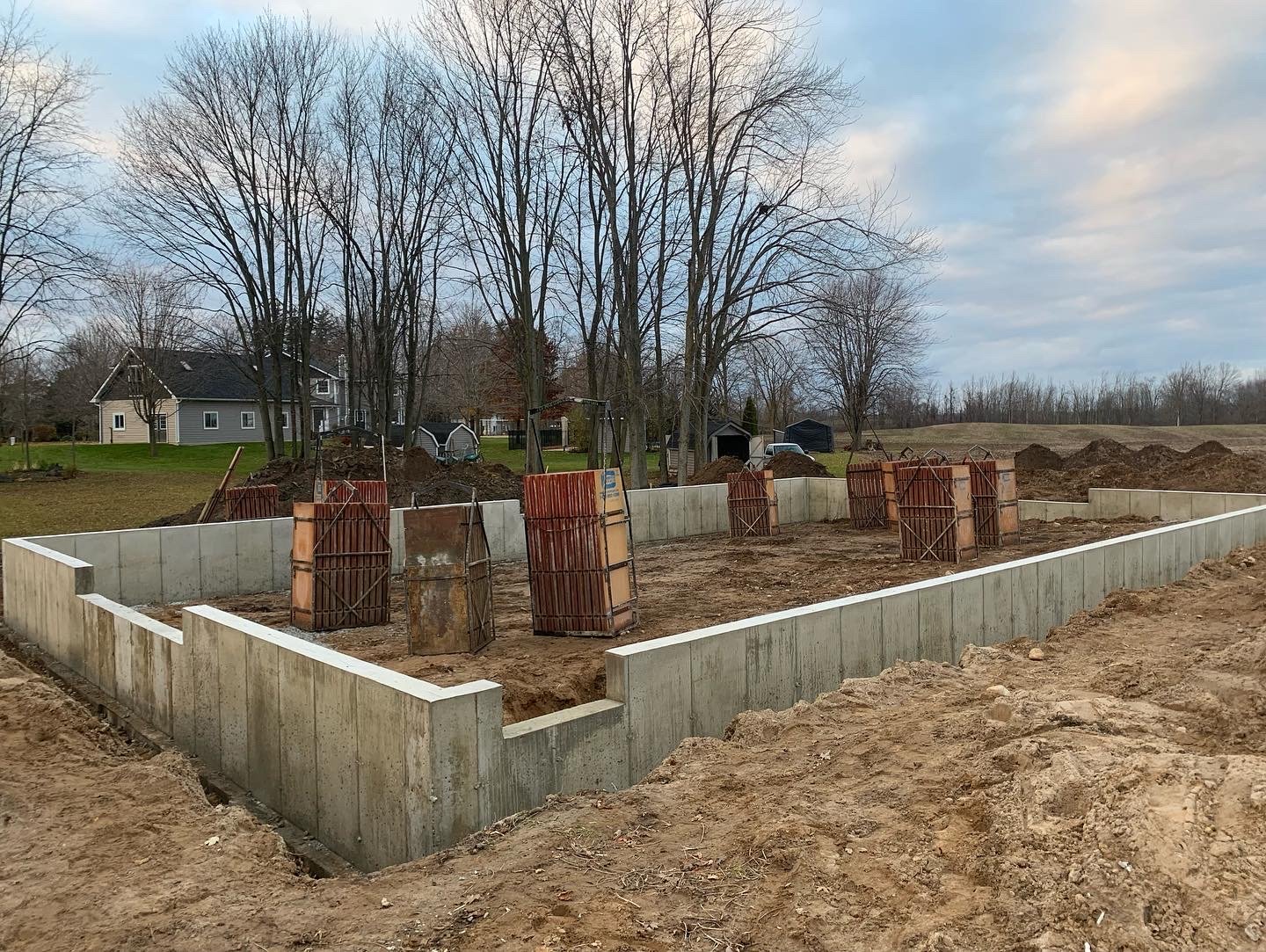 Construction site with concrete foundation walls and wood formings for columns, dirt and piles of soil, set in a residential area with trees and houses in the background.